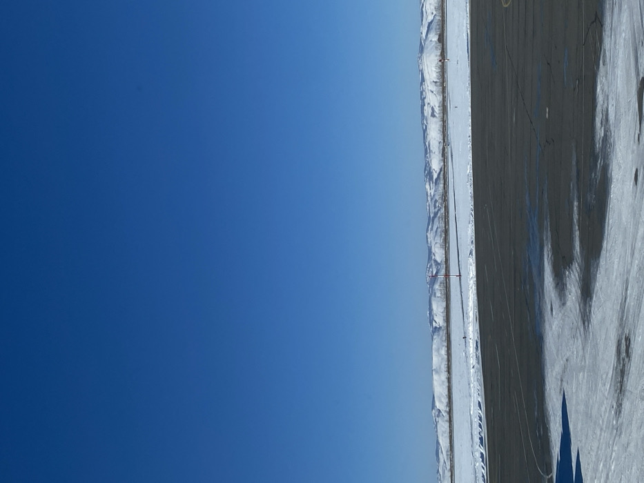 The image, when properly oriented (rotated 90 degrees counter-clockwise), displays a wide-angle landscape primarily composed of a vast blue sky, distant snow-capped mountains, and a flat, dark foreground.
The upper portion of the image is dominated by a clear, deep blue sky, which fades slightly to a lighter hue closer to the horizon. There are no clouds visible, indicating a bright and sunny day.
Stretching across the midground, just below the sky, is a long, undulating range of snow-capped mountains or large hills. These mountains appear distant and rugged, their white peaks and slopes contrasting sharply with the deep blue sky.
The lower half of the image features a large, flat, dark-colored ground surface, resembling an asphalt runway, a vast paved area, or a large parking lot. This surface shows visible cracks and is partially covered with patches of snow and ice, suggesting cold or wintery conditions. Long, distinct dark shadows are cast across this paved area, extending from the right side of the frame towards the left, indicating bright sunlight likely from a low angle, such as morning or late afternoon. The shapes of these shadows suggest they might be cast by structures or objects just outside the frame.
Between the dark foreground and the distant mountains, there is a narrower strip of lighter, snow-covered ground, which could be a cleared path, road, or simply a flatter expanse of snow. Within this strip, two slender poles, each topped with a small red flag or marker, are sparsely visible, adding minor points of interest to the otherwise desolate scene.
The overall impression is one of a vast, open, and cold environment with clear weather. There are no people, animals, or significant man-made structures (apart from the paved surface and small poles) visible in the frame, conveying a sense of isolation and natural expanse. No text is visible in the image.