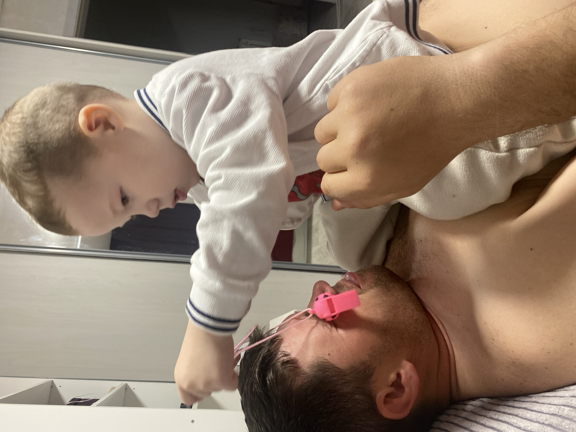 A young child, dressed in a white, long-sleeved shirt with blue trim, is standing over a man lying on his back. The man is shirtless and has a pink whistle on a string near his face. The environment appears to be indoors, possibly a bedroom, with a white cabinet in the background. The city of Sochi, Russia, is not discernible from the image.