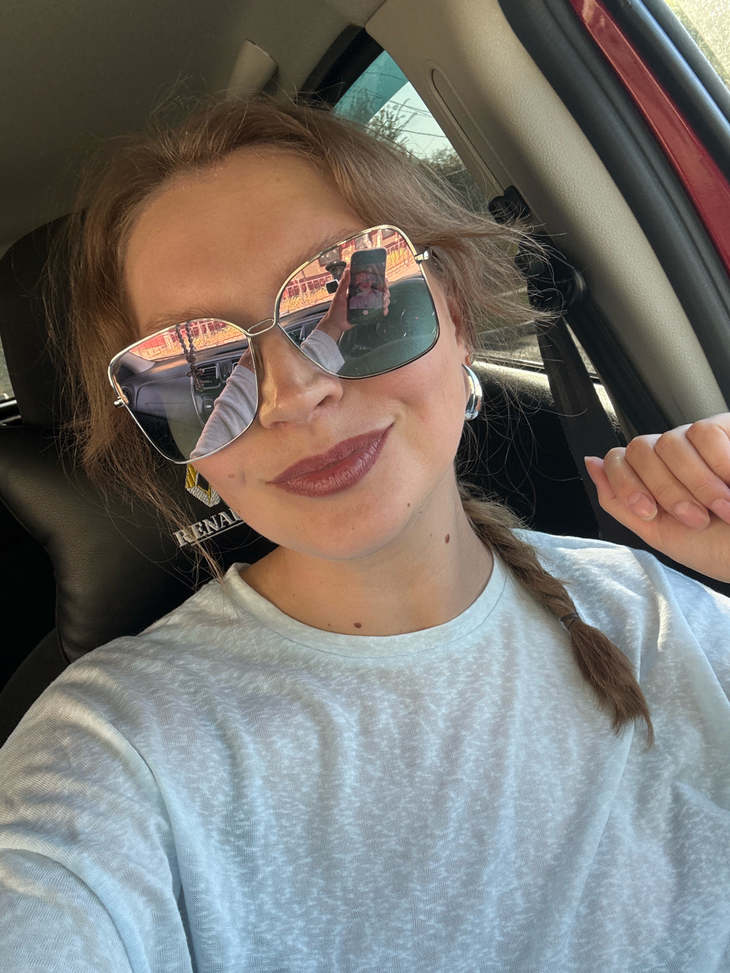 A young woman with blonde hair in a braid, wearing a light blue t-shirt and large sunglasses, takes a selfie in a car. Her reflection in the sunglasses shows her holding a phone and the car's interior, including a steering wheel and dashboard. A small mole is visible on her neck, and another on her chest. The car's interior includes black leather seats and a red exterior panel is visible through the window. The background outside the window shows trees and a power line. The emblem of "Renault" is visible on the headrest of the driver's seat. The photo was taken in Aksay, Kazakhstan.