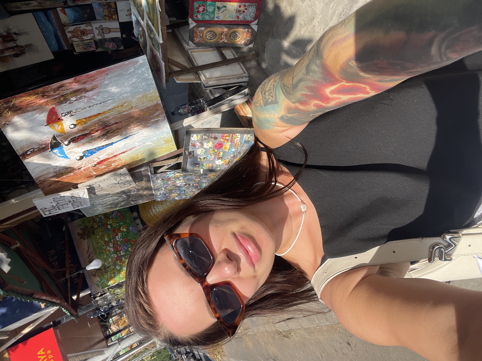 A woman is taking a selfie outdoors with a bright sun, casting shadows. She is wearing sunglasses and a black top, with a white purse strap visible. Her left arm, adorned with a colorful tattoo, is extended towards the camera. Behind her, an array of paintings and small decorative items are displayed, suggesting a market or souvenir stall. One painting prominently features three sailboats with colorful umbrellas. The ground appears to be paved. The overall scene suggests a sunny day in a bustling outdoor market, possibly in Ubud District, Indonesia, given the context.