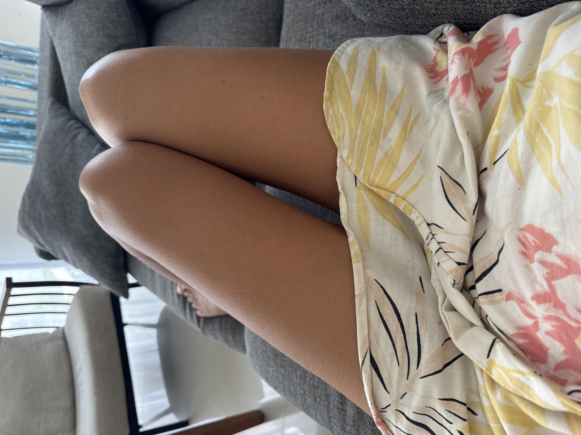The image depicts a person's legs resting on a gray couch. The person is wearing a floral print dress or shirt. The background includes a glimpse of a window and a chair. The exact location within Kuta Selatan, Indonesia cannot be determined from the image. There are no actions, interactions, or people, other than the person's legs.