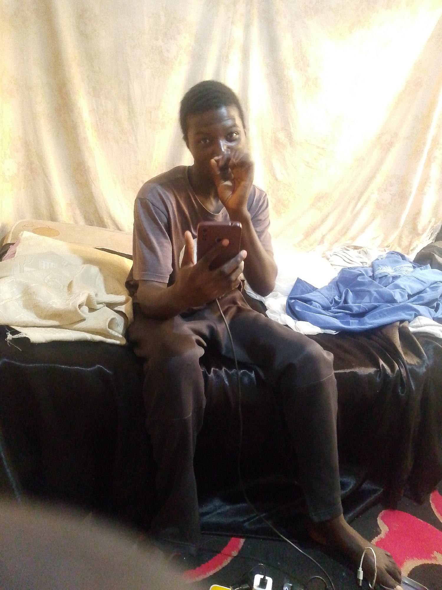 A young Black male, seated on a bed, is holding a smartphone in his right hand. He is looking towards the viewer and has his left hand near his mouth, as if holding something to it. He is wearing a dark-colored t-shirt and dark pants. The bed is covered with dark fabric, and to the left and right are piles of fabric, one appearing to be a light-colored patterned cloth and the other a blue sheet. Behind him, a light-colored curtain hangs, with a bright light source behind it. The floor, partially visible at the bottom of the image, appears to be carpeted with a patterned design. The scene is indoors, likely a bedroom, and the overall lighting is dim and artificial. There are no visible indicators to suggest the specific location within Jos, Nigeria.