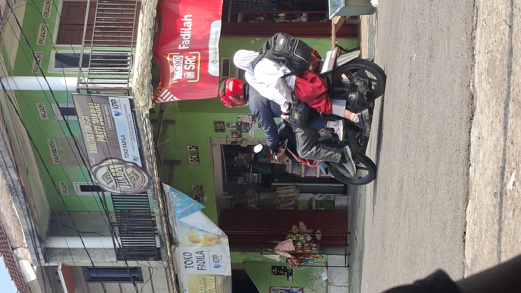 A man wearing a dark blue jacket and a red helmet is riding a motorcycle. A person, likely a child, wearing a white headscarf and a red uniform, is sitting behind the man on the motorcycle. The person behind is carrying a black backpack. They are on a street in front of a building with green walls, a balcony, and several shops. A banner on the building reads "KUALITAS TINGGI PASTI JUARA!" and another banner on the right side of the building has "TOKO SRC Fadilah" written on it. The street appears to be paved with asphalt. The scene is likely in Cilaku, Indonesia, given the context of the businesses.