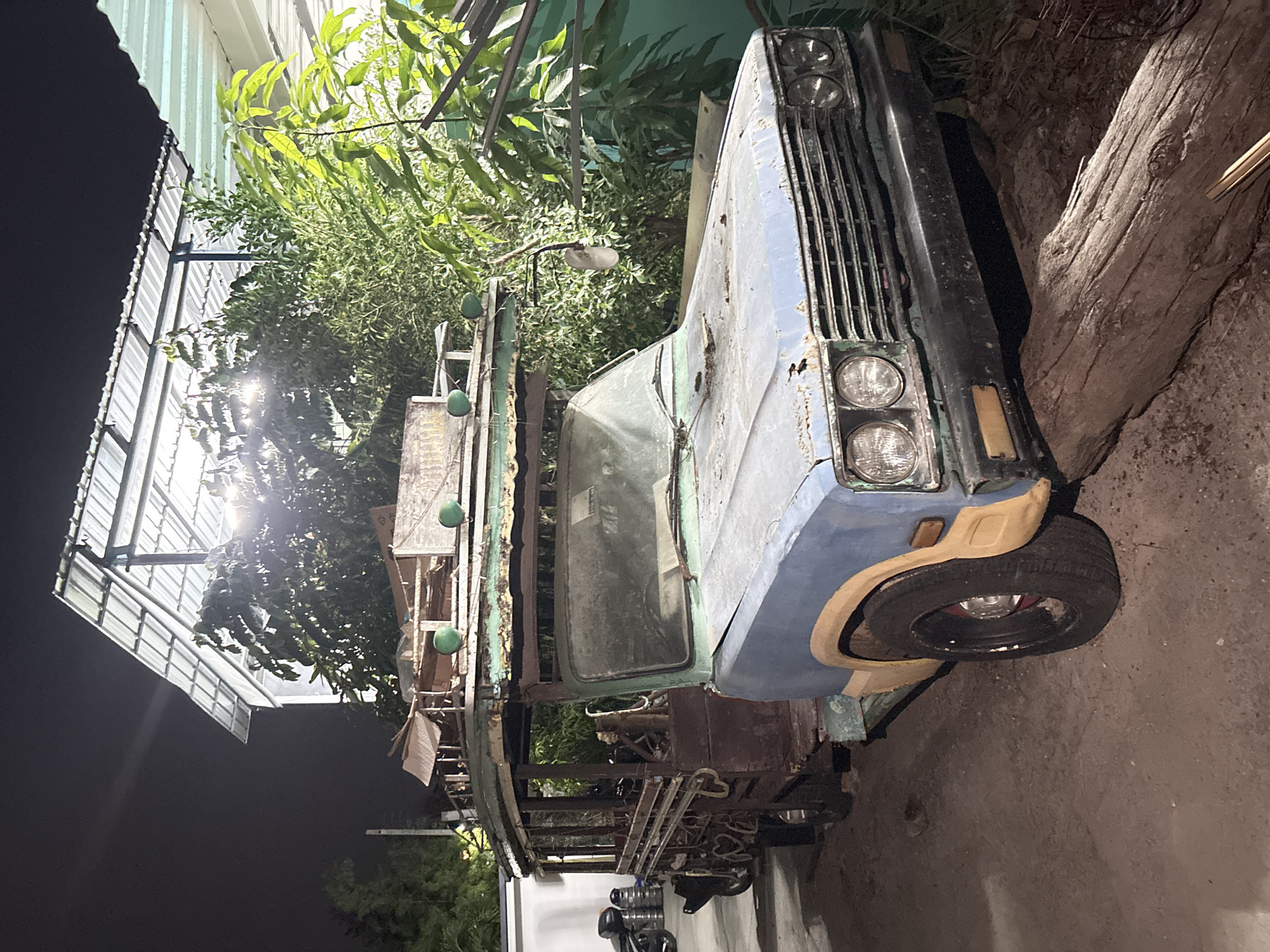 An old, dilapidated blue and grey truck, partially obscured by foliage, is parked beside a tree trunk in Baan Bang Rak, Thailand. The truck has a rusty grille, a partially visible cab interior, and a roof rack. Miscellaneous objects are underneath the truck. The scene is lit by artificial lights from the left, while the background fades into darkness.