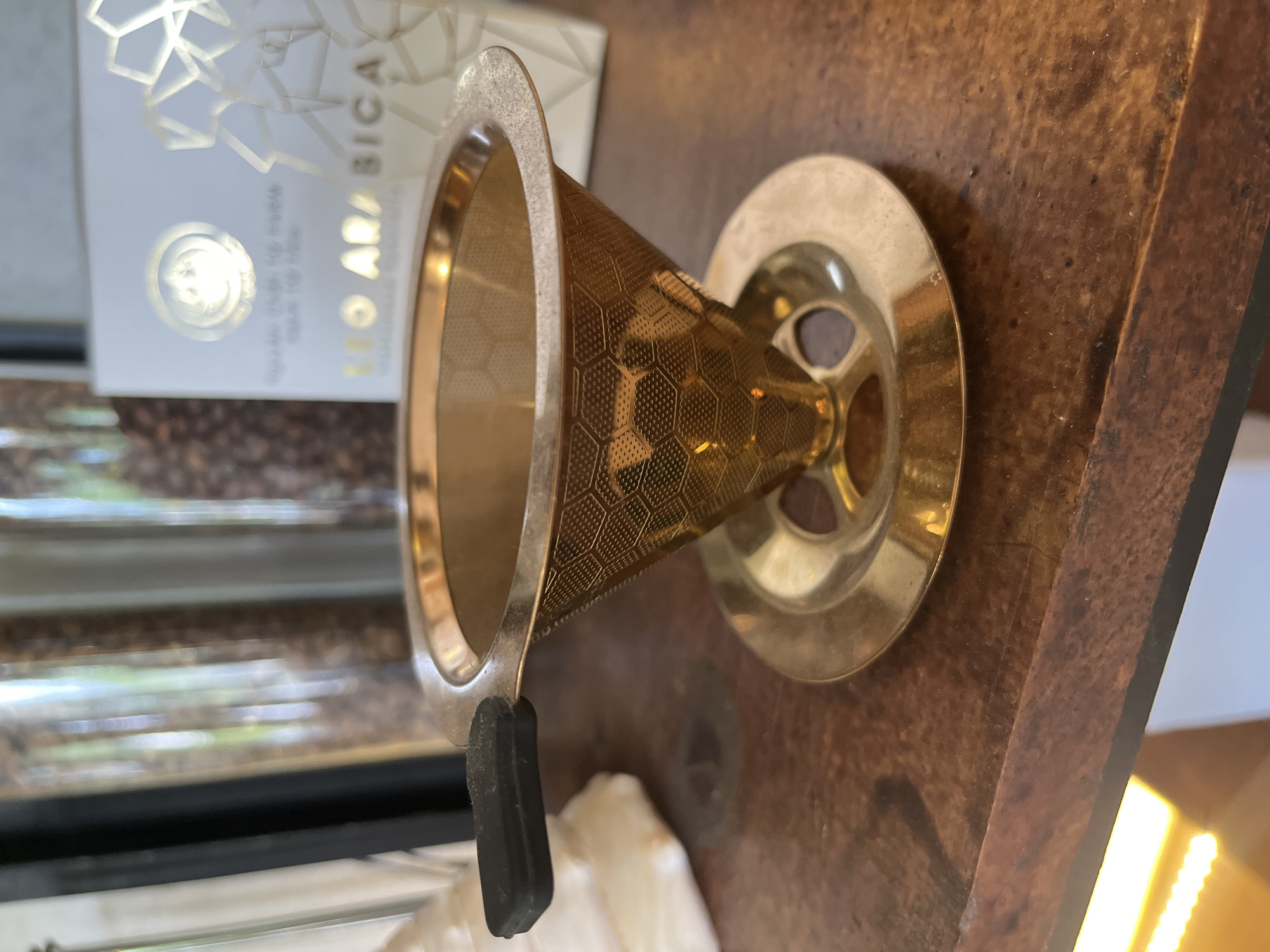 The image depicts a gold-colored metal coffee filter sitting on a dark brown surface. The filter is cone-shaped, with a wider circular opening at the top and a smaller, perforated cone below. It has a wide, flat base with three triangular openings. A rectangular black rubber handle extends from the side of the filter. Behind the filter, there is a white cardboard box with text in a foreign language ("LEO ARABICA"). Below the box, a blurry background suggests a coffee bean display behind glass. The surface appears to be a wooden table or countertop with a visible grain. Light can be seen in the background coming from the lower right corner, suggesting a window. The location, Bac Nha Trang Ward, Vietnam, cannot be verified from the image alone.