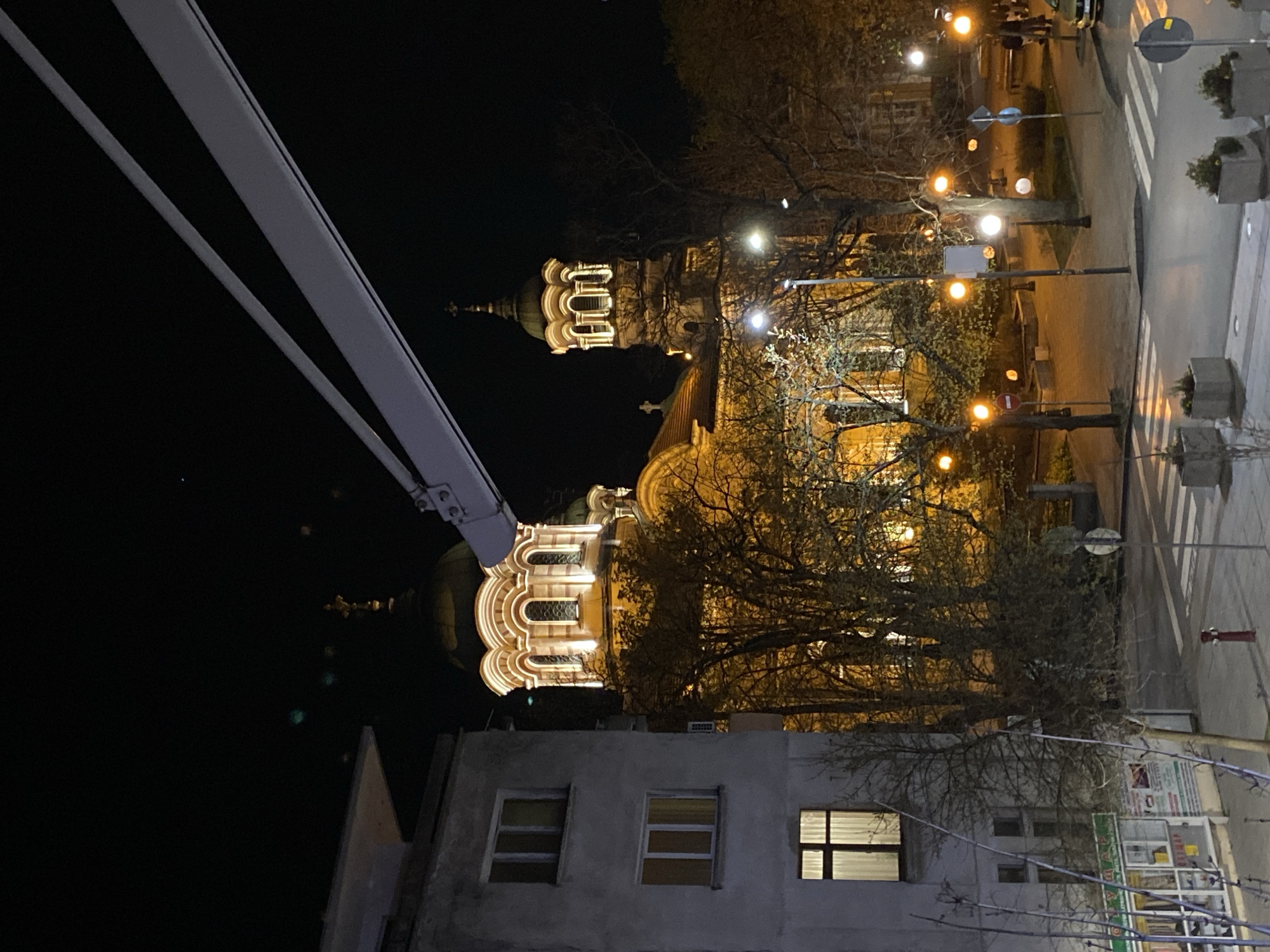 The image shows a nighttime view of the Sveti Dimitar Church in Stara Zagora, Bulgaria. The church is illuminated, and its architectural details are visible. A building is in the foreground with windows. Street lights are present along the street, and a crosswalk is visible. Bare trees are scattered throughout the scene. A structure or equipment is visible at the top-left corner, which could be part of a building or a construction element.