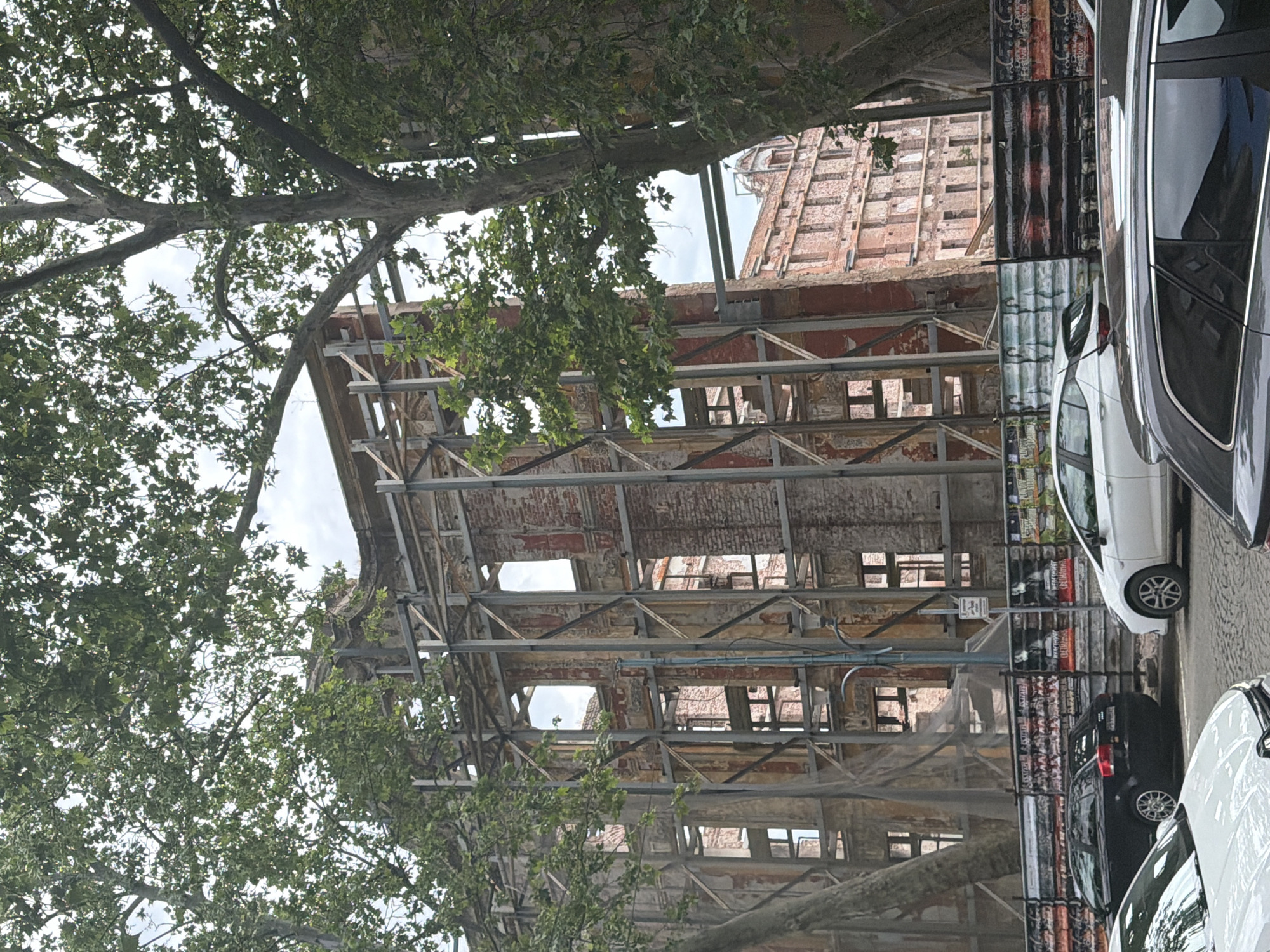 The image captures the partial ruins of the Small Basilica in Plovdiv, Bulgaria, framed by trees on the left. The basilica's skeletal structure is visible, with a mix of exposed brickwork and metal scaffolding supporting the crumbling walls. A portion of what appears to be the roof structure is also present, showing the extent of the damage. In the background, the top of another brick structure is visible.
To the right of the basilica, a black metal fence or barrier stands, and cars are parked along the street. The sky is overcast, providing a soft, diffuse light. The overall impression is one of historical decay juxtaposed with the modern urban environment.