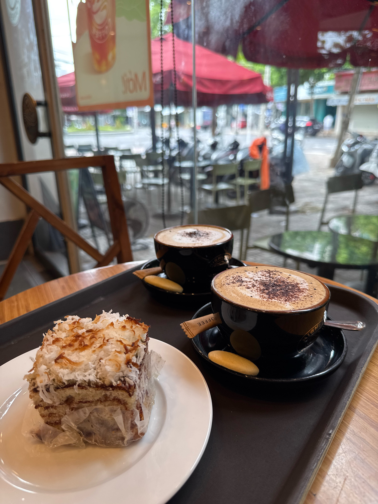 This image depicts a close-up of a tray with two cups of coffee and a slice of cake, set against a blurry backdrop of an outdoor cafe in Nha Trang, Vietnam. The tray, made of dark material, holds a white plate with a piece of cake topped with shredded coconut. To the right, two black coffee cups with saucers are visible, each containing a cappuccino with cocoa powder dusting. A small biscuit rests in one saucer, and a packet of sugar with the word "COFFEE" is tucked into the other. The background reveals the exterior of the cafe, with red awnings, tables, and chairs, and a street scene with motorbikes and buildings characteristic of Nha Trang. The overall impression is of a casual coffee break at an establishment in this Vietnamese city.
