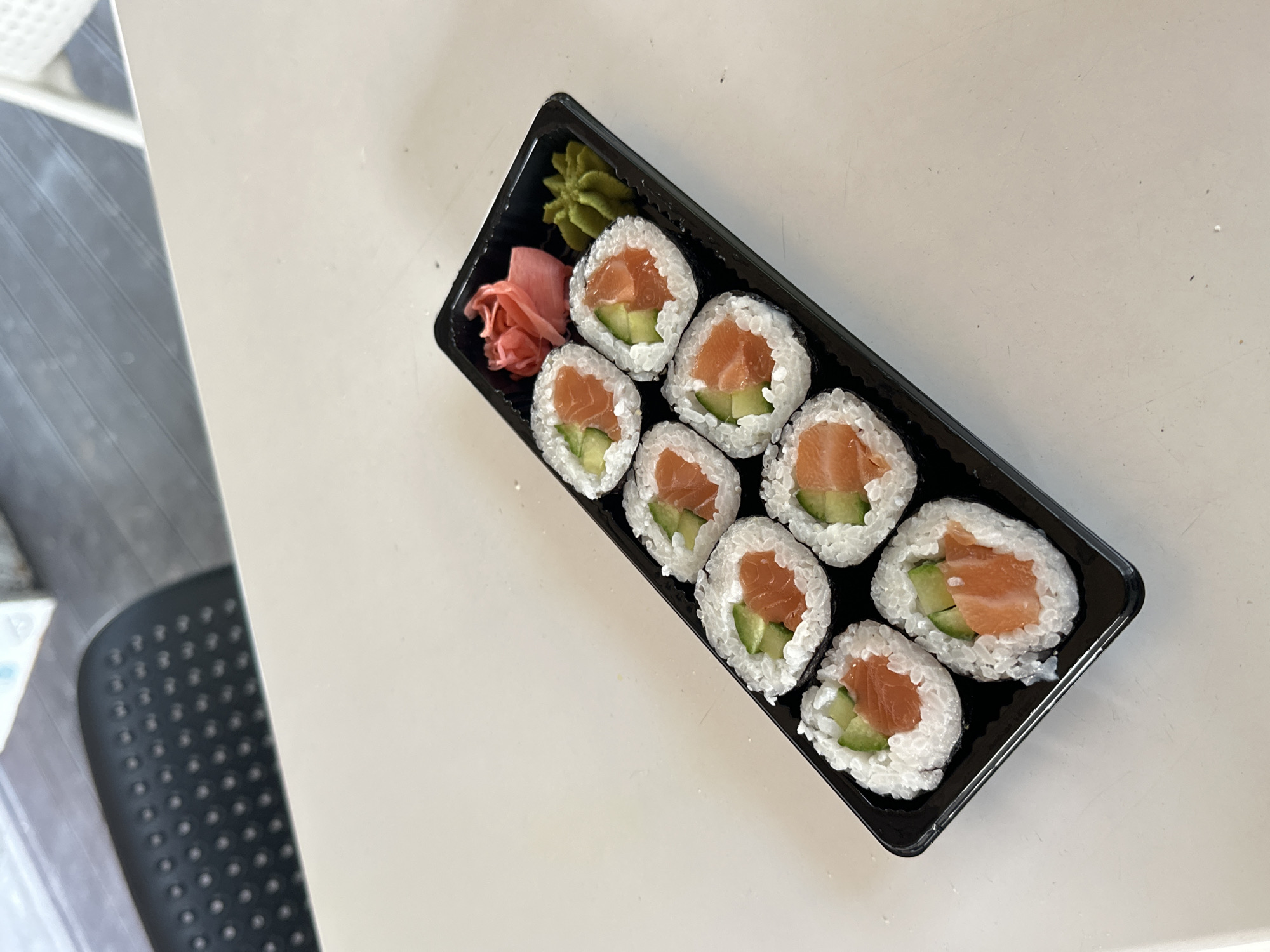 The image shows a black plastic container of salmon sushi rolls, along with wasabi and pickled ginger, resting on a white surface. The rolls contain salmon and cucumber. There are no people visible. The location is not identifiable as Varna, Bulgaria.