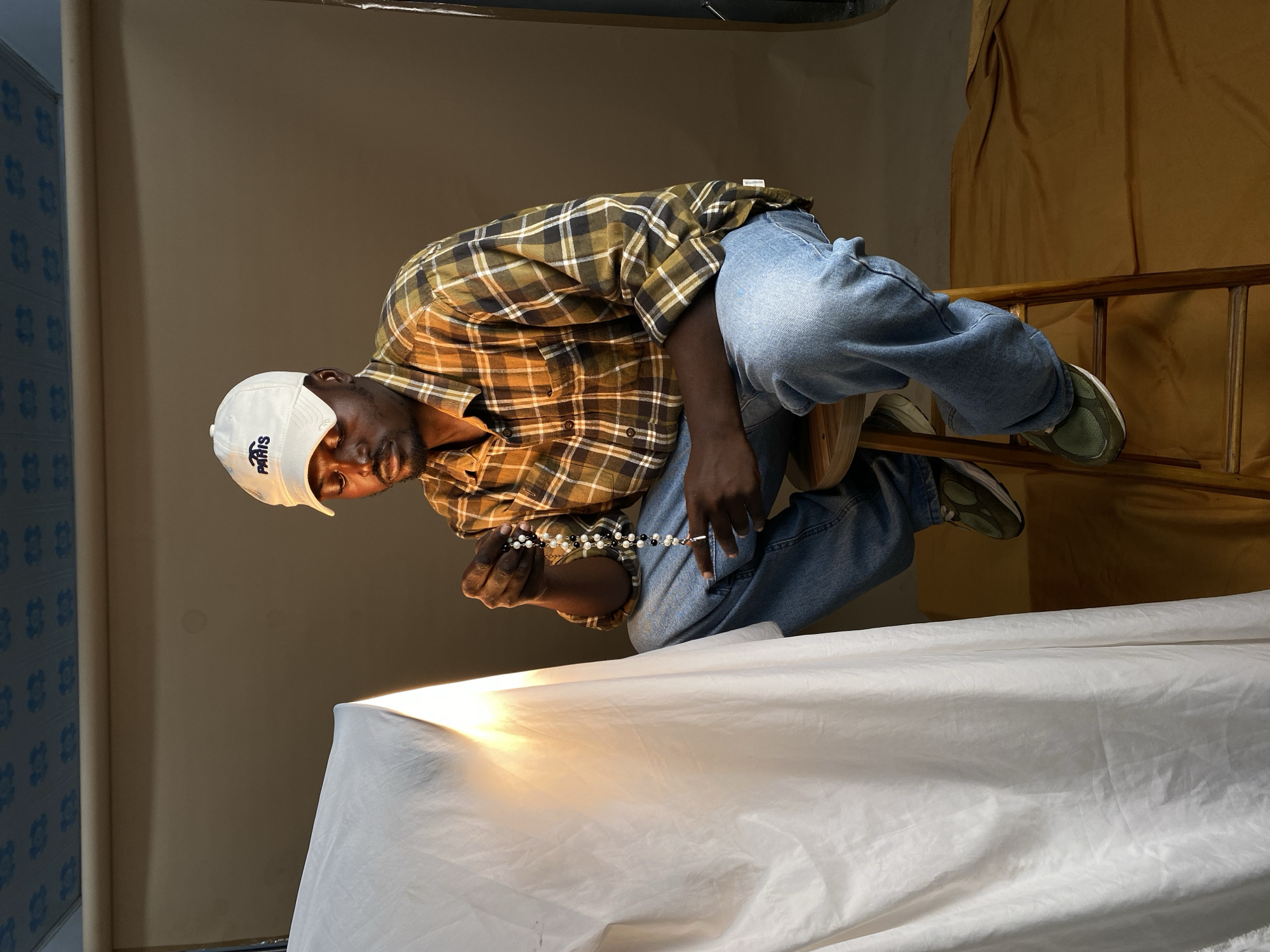 A Black man wearing a white baseball cap with "PARIS" embroidered on it, a plaid long-sleeved shirt in shades of brown and olive, and light-wash blue jeans is sitting on a wooden stool in what appears to be a studio setting. He is holding a string of black and white beads in his hands, looking down at them. To his left, a large piece of white fabric is draped and lit, casting a bright glow. The background is a neutral brown. The floor is covered with a yellowish-brown fabric. The ceiling is visible with a patterned design. The scene is located in Gura Top, Nigeria.
