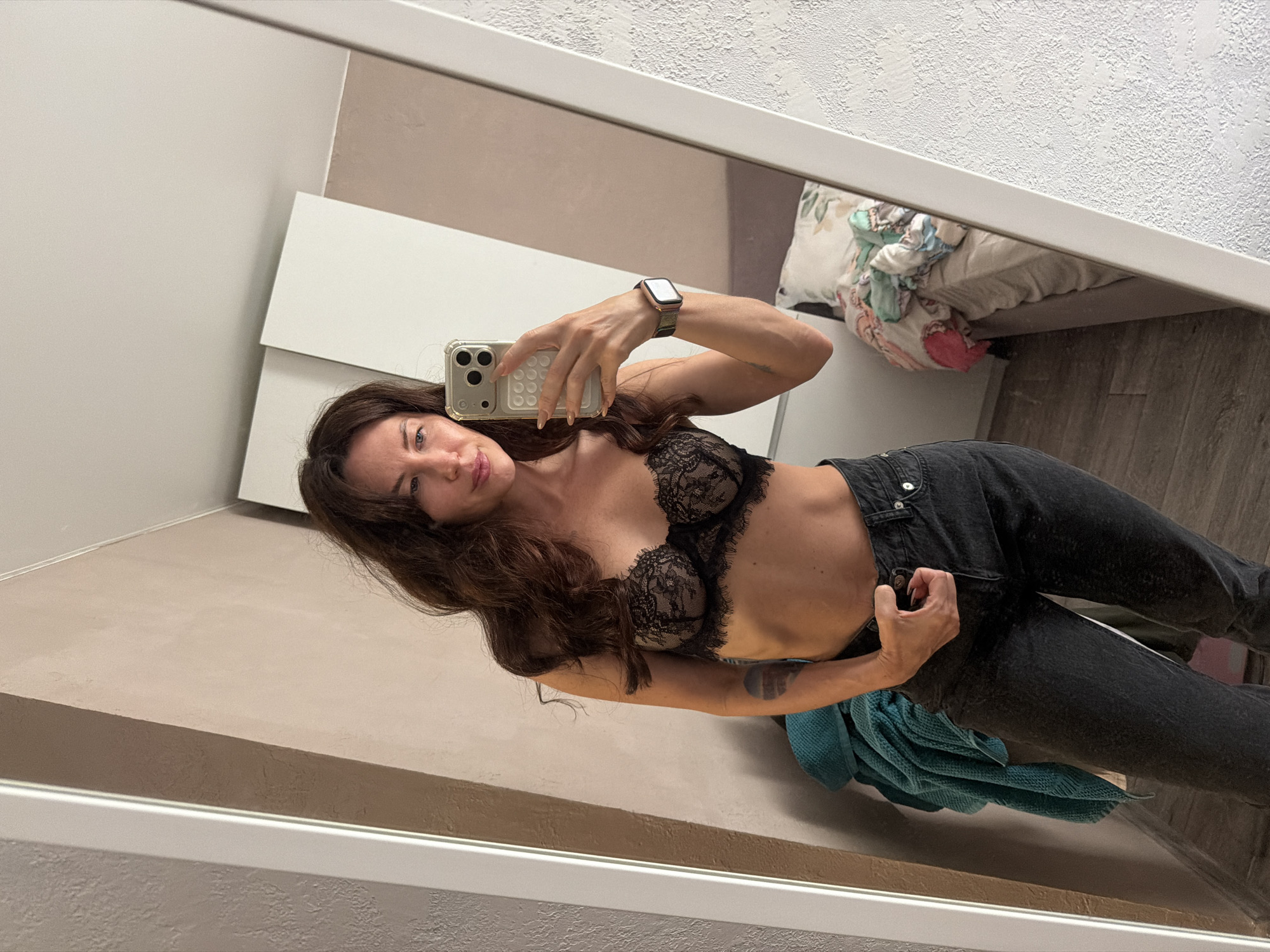 A woman with long, dark hair is taking a selfie in a mirror. She is wearing a black lace bra and dark wash jeans. Her left arm is resting on her hip, and her right hand is holding her phone, which she is using to take the photo. A smartwatch is visible on her right wrist. Behind her, a blue towel is draped over a railing, and a bed with colorful bedding is visible in the reflection. The room appears to be a bedroom, with a closet door in the background. The image is taken in a well-lit room with neutral-colored walls. There is no information to suggest the location is in the city of Perm, Russia.
