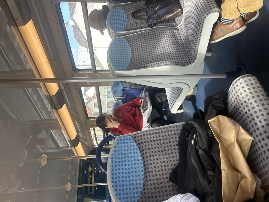 The image depicts the interior of a public transport vehicle, likely a train or tram, located in Villemomble, France. Three individuals are visible within the cabin. In the mid-ground, a woman with dark hair, wearing a red long-sleeved top and dark pants, is seated. She holds a smartphone in her right hand and has her left hand pressed against her forehead. A white earbud is visible in her right ear. A grey blanket or garment rests on her lap, and a blue tote bag occupies the seat next to her. Further back and to the right, the rear view of a person wearing a brown baseball cap and dark clothing is visible; a dark brown leather shoulder bag hangs on the seat directly in front of them. In the immediate foreground, the lower legs and feet of a third passenger are shown, dressed in tan pants, striped socks, and brown shoes. A black garment and a crumpled brown paper bag are placed on the seat in front of this foreground passenger. The vehicle's interior features patterned grey and blue seating, overhead fluorescent lighting, and vertical support poles. Through the windows, residential buildings with pitched roofs and a clear blue sky are visible, indicating an urban or suburban setting.