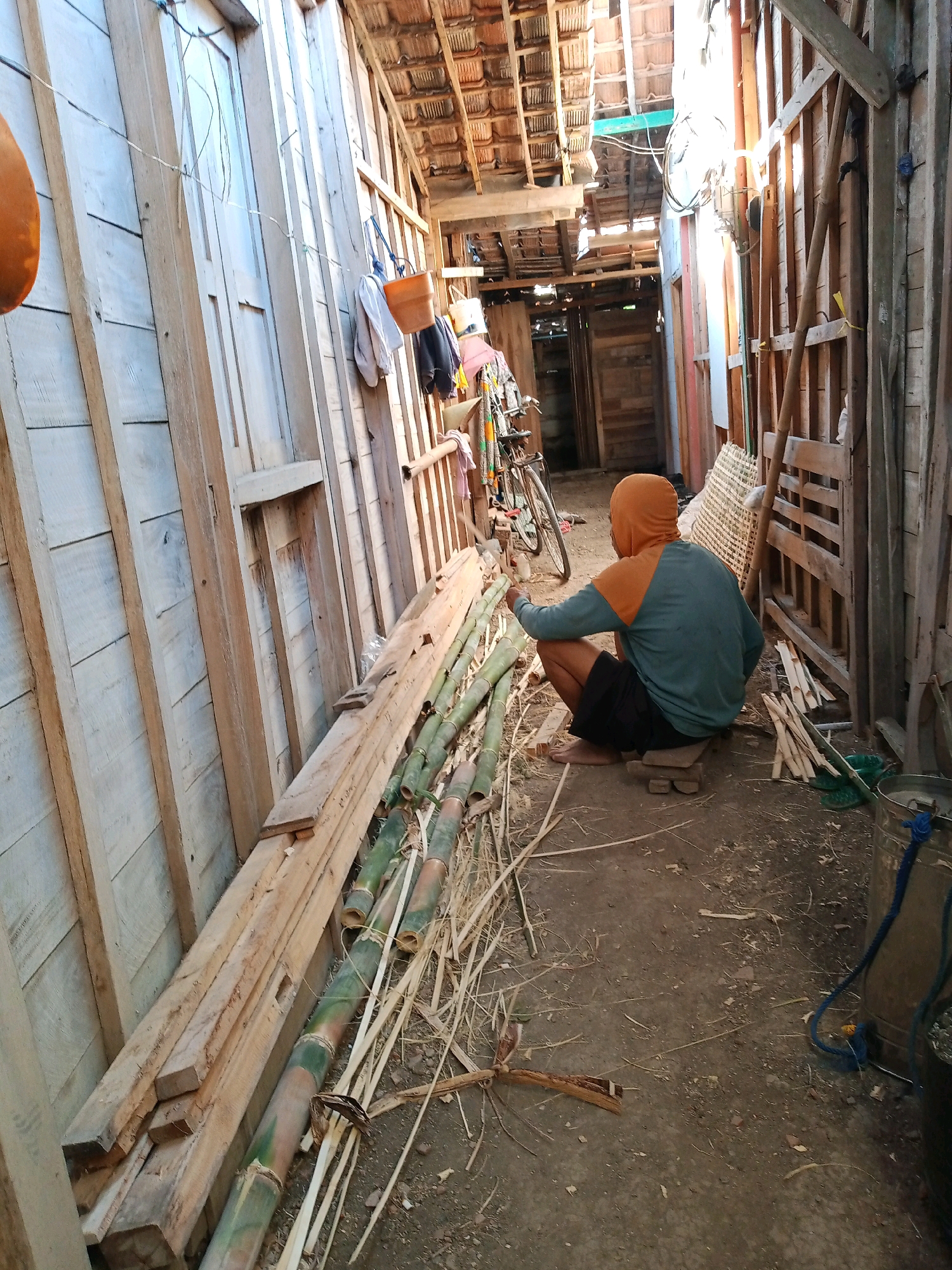 The image depicts an outdoor scene in Kemiri, Indonesia, likely a workshop or storage area for building materials. In the foreground, a person wearing a hooded sweatshirt and shorts is squatting on a wooden plank. The person is facing away from the camera, with their left arm extended towards a pile of bamboo poles. To the left of the person, a stack of lumber is leaning against a wooden wall. The wall appears to be constructed from rough-hewn wooden planks. Beyond the person and the bamboo, a narrow pathway leads into a dimly lit structure with a tiled roof. A bicycle is visible in the background, leaning against the wall. The ground is covered in dirt and debris, with scattered pieces of wood and dried vegetation. The overall impression is of a rustic, utilitarian environment, possibly associated with construction or craftwork.