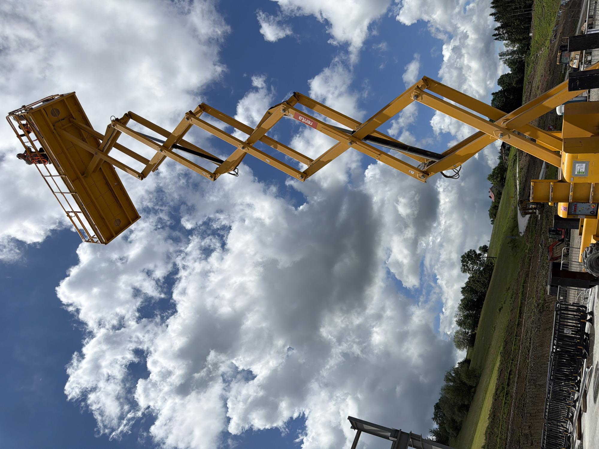 A yellow scissor lift, raised high into a partly cloudy blue sky, is the primary subject. Two individuals are visible in the lift's platform at the top. The scissor lift's complex geometric structure is prominent, extending upwards from a base unit on the ground. The surrounding environment appears to be a construction site or outdoor event setup. In the background, a green, hilly landscape with trees and some buildings can be seen. Fencing and other construction equipment are present on the ground level. The image appears to be taken on a bright, possibly sunny day, with dramatic cloud formations. The specific location of Janowice, Poland is not discernible from the visual information within the image itself.