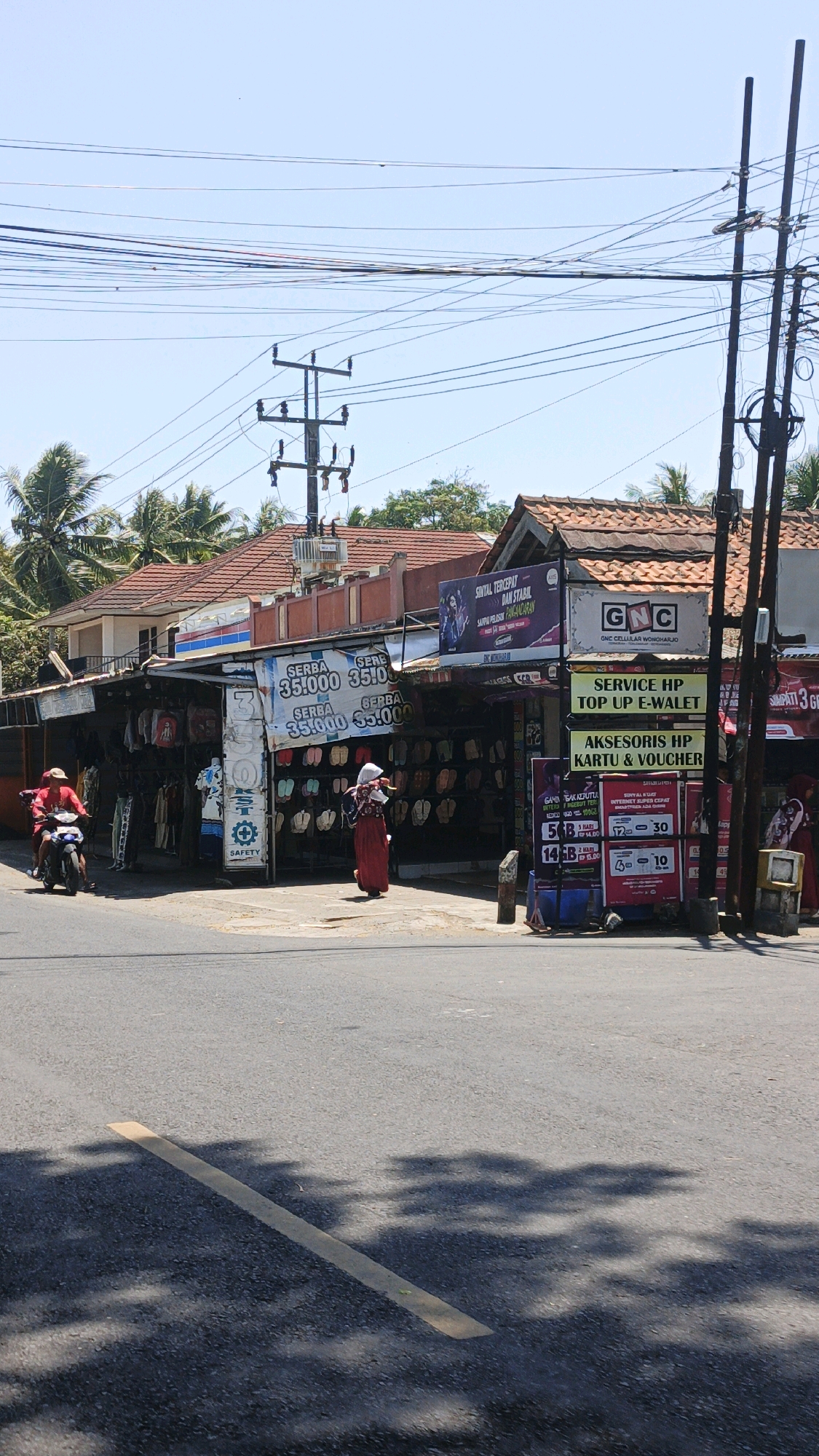 The image shows a street scene in Desa Wonoharjo, Indonesia, on a sunny day. In the foreground, a grey asphalt road has a faded yellow line marking a lane or boundary. Shadows of trees are cast across the road. Behind the road, a row of shops with open fronts displays merchandise, including clothing and footwear, likely sandals. A man in a red shirt is riding a blue motorcycle and appears to be turning or stopped at the intersection. A woman in a long red dress and white head covering walks along the sidewalk in front of the shops. Another woman in a similar red dress and white head covering is visible further down the street, partially obscured. Several signs are posted on the buildings, with some in Indonesian. One prominent sign advertises "GNC CELLULAR WONOHARJO". Overhead, a complex network of electrical wires and utility poles spans the sky. The buildings appear to be single-story structures with red tiled roofs. The overall atmosphere is that of a bustling local market or commercial area.
