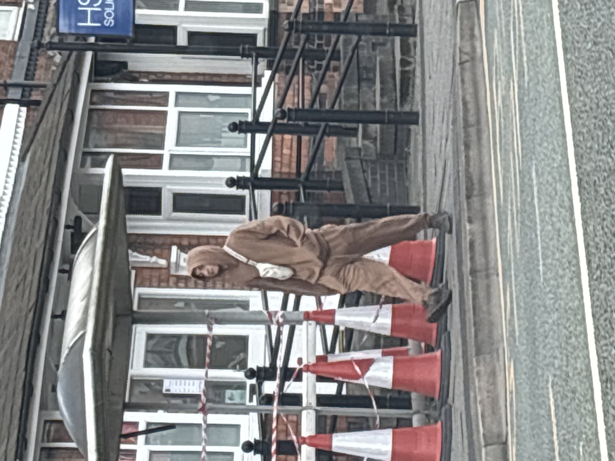 A man wearing a brown hooded jumpsuit and a white shoulder bag is lying horizontally across a row of orange and white traffic cones on a pavement. The pavement is adjacent to a road with white line markings. Behind the cones, there is a building with multiple windows, a black metal railing, and a blue sign that says "HS" in white lettering. The location is Crewe, United Kingdom.