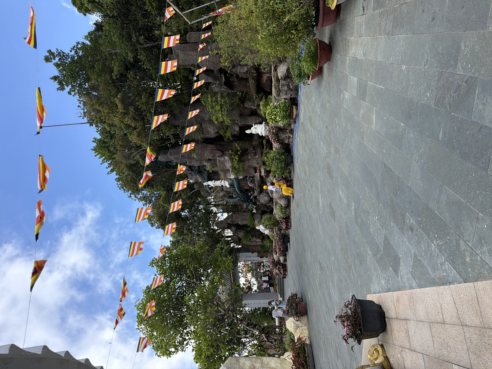 The image depicts a courtyard area in front of a Buddhist temple, likely located in the city of Đà Nẵng, Vietnam, given the cultural context. The foreground is dominated by a paved stone courtyard. To the left, a section of a building with a textured white facade and an archway leading into the temple grounds is visible. Beyond the archway, several people are seen walking or standing.
The central and background areas feature a man-made rock garden or cliff formation with a small waterfall. Several Buddha statues are placed within this rock garden, including a prominent white statue of a seated Buddha. A decorative dragon sculpture is also present, partially obscured by foliage.
A person wearing a yellow outfit is seated on a step near the rock garden. Various potted plants and flower beds with pink and red flowers are strategically placed around the courtyard and the rock garden, adding color and life to the scene.
Numerous Buddhist prayer flags, in vibrant colors such as yellow, red, white, and blue, are strung across the courtyard at different heights, fluttering in the breeze under a bright blue sky with scattered clouds. The flags suggest a religious or celebratory occasion.
The overall atmosphere appears serene and spiritual, with a blend of natural elements, religious iconography, and architectural features typical of a Vietnamese Buddhist temple complex.