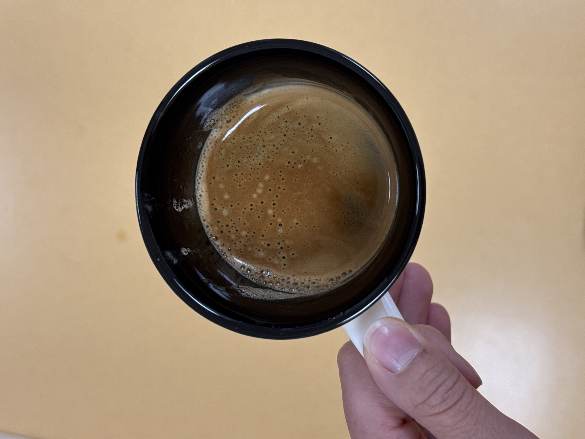 A hand is holding a black mug filled with dark brown espresso. The espresso has a light brown crema with small bubbles on the surface. The mug has a white handle. The background is a plain, light brown surface. This image was taken in Pleven, Bulgaria.
