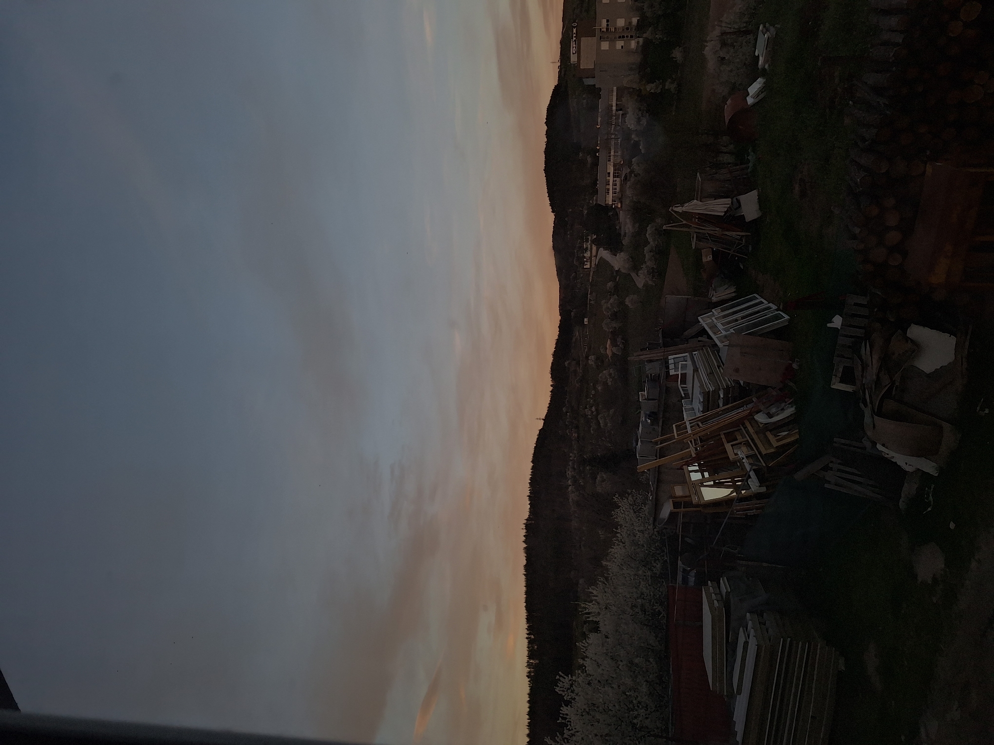 The image shows a dusky landscape, possibly taken at dawn or dusk. In the background, there is a silhouette of a forested hillside. Closer to the foreground, there is a cluttered area with what appears to be discarded construction materials, including window frames and lumber. The sky is multi-toned, with a mix of blues and pinks. Buildings are visible atop the forested hill.
Without further context, positively confirming that this location is in Panagyurishte, Bulgaria is not possible, although the urbanized buildings combined with the surrounding forests would be consistent with such a location.