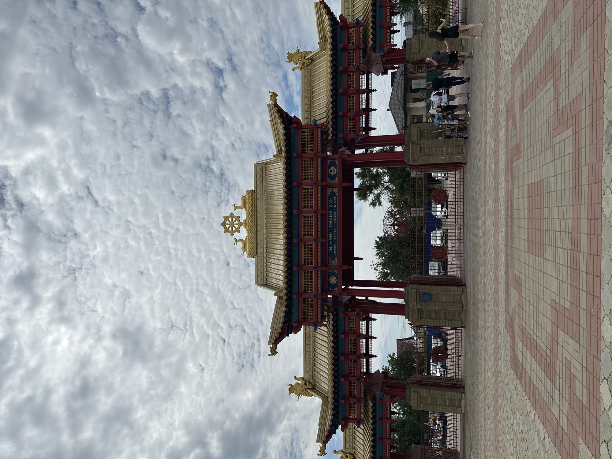 The image depicts the entrance gate to the "Golden Abode of Buddha Shakyamuni" temple in Elista, Russia. The gate is a large, ornate structure with traditional Tibetan Buddhist architectural elements. It features multiple tiers of golden roofs, red pillars, and intricate decorative carvings, including golden dragon motifs and a Dharma wheel at the apex. The gate is flanked by brick pillars and intricate metal fencing. Through the gate, a glimpse of the temple grounds and surrounding trees can be seen, along with several parked cars. In the foreground, a paved courtyard with a red and beige patterned brickwork leads to the gate. A group of people, appearing to be tourists or visitors, are gathered near the entrance, some interacting with each other and others looking towards the temple. The sky above is overcast with scattered clouds, suggesting a temperate day.