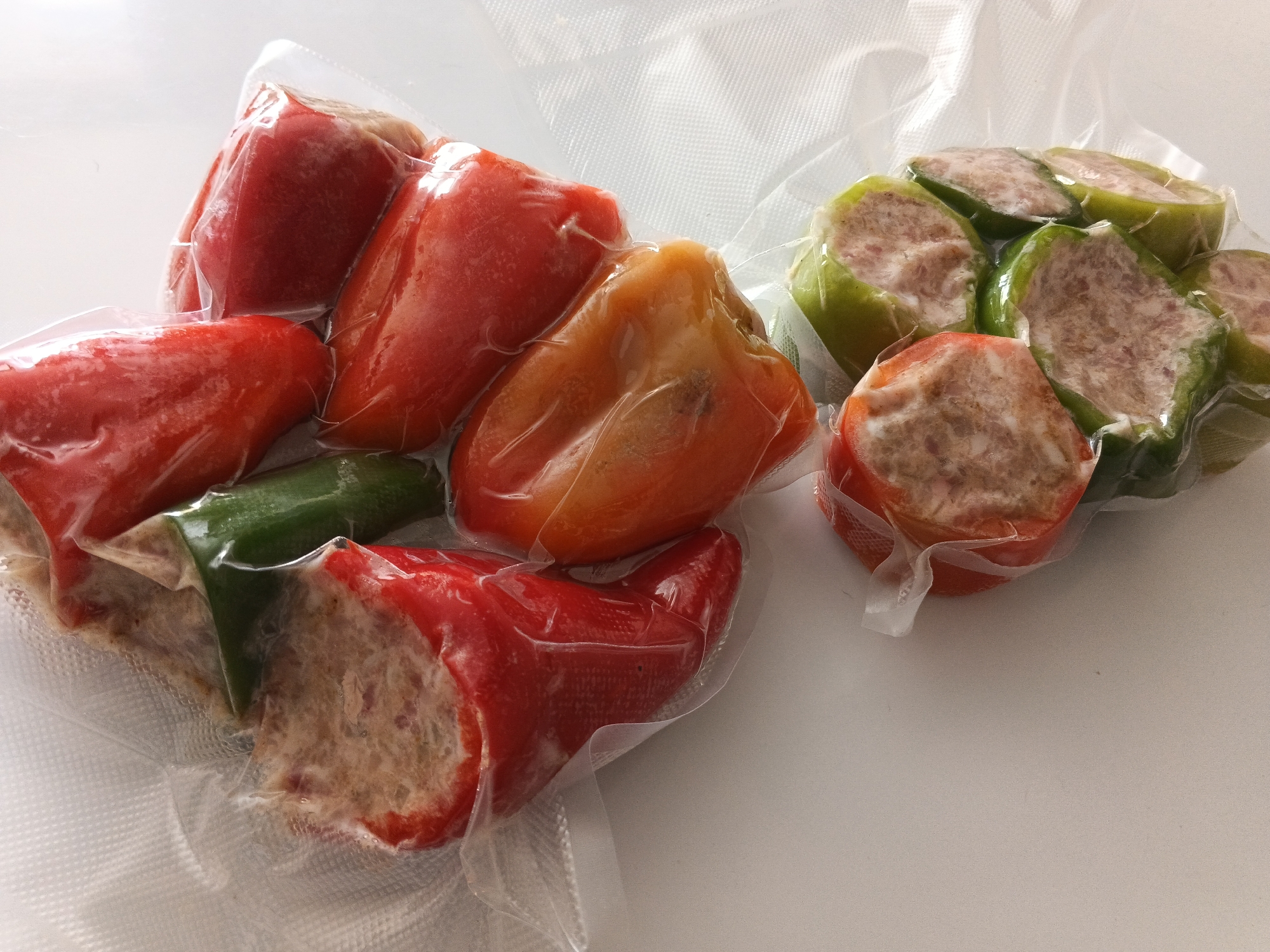 Two vacuum-sealed packages of stuffed bell peppers are displayed on a white surface. The package on the left contains red and orange bell peppers, while the package on the right contains green bell peppers. The peppers are halved and filled with a mixture of ground meat and seasonings. The background is a plain white surface. This image does not provide any information about the city of Металлистов, Russia.