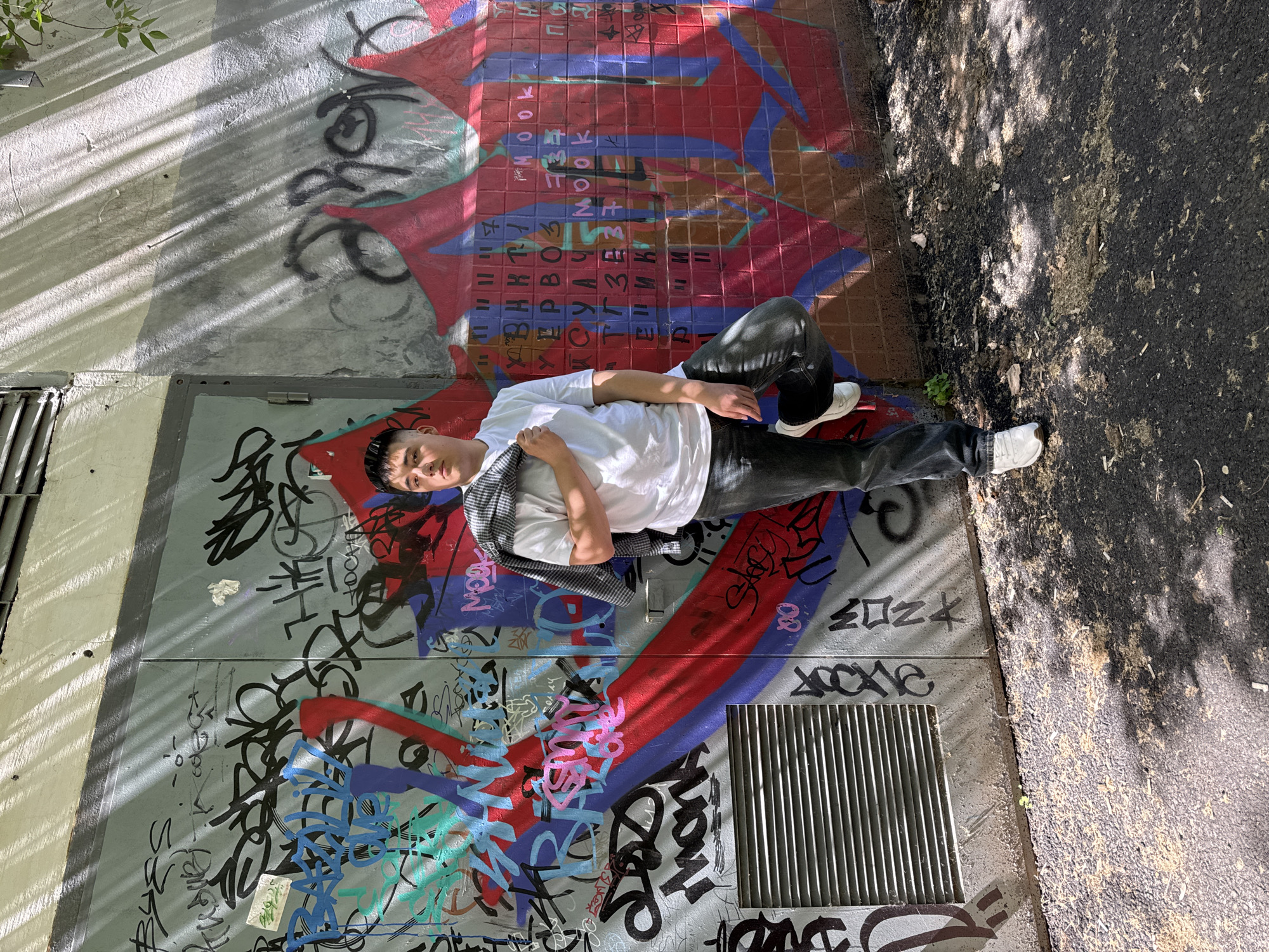 A young man is lying on a graffitied wall next to a street in what appears to be an urban setting. The wall features layers of graffiti in various colors, including red, blue, black, and white. Some of the graffiti includes Cyrillic script, possibly related to the location of Chaikovsky, Russia. The man is wearing a white t-shirt, dark jeans, and white sneakers. He has a plaid shirt or jacket draped over his body. The environment includes sunlight filtering through trees, casting shadows on the wall. Part of the street or sidewalk is visible adjacent to the wall.