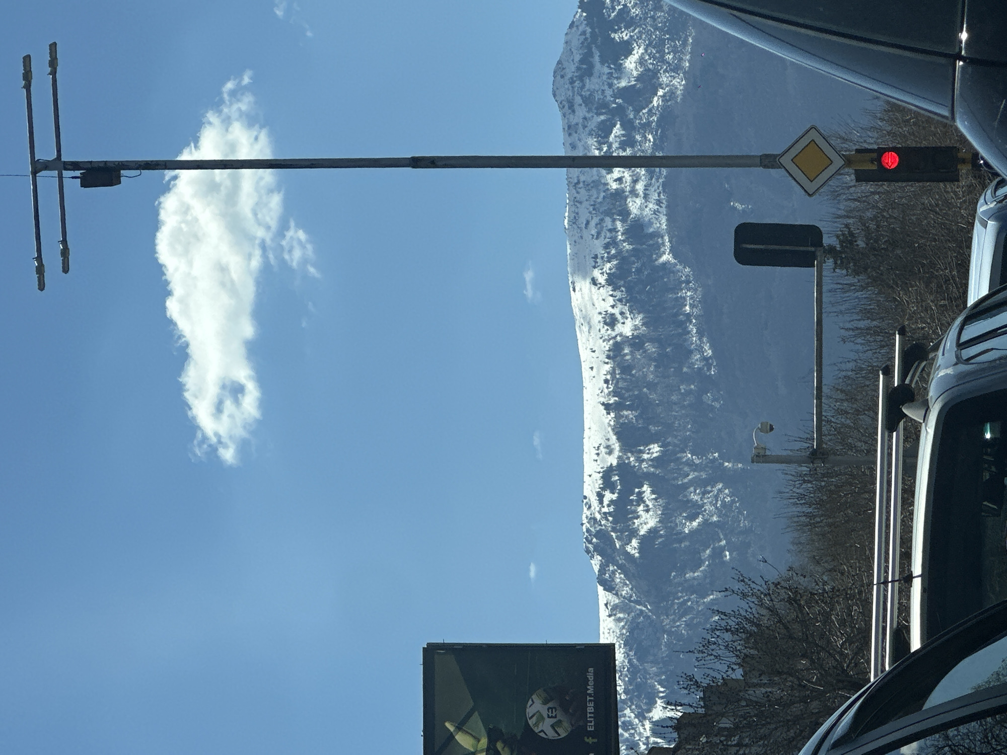The image, oriented sideways, depicts a view from a vehicle, possibly in Sofia, Bulgaria, based on the presence of an "ELITBET.Media" sign which indicates that. A snow-covered mountain is visible in the background against a blue sky with a single prominent cloud. The foreground includes traffic infrastructure such as an antenna structure, a yellow diamond-shaped traffic sign, and a traffic light showing a red light. Cars and bare tree branches are visible, obscuring some of the foreground. A billboard is partially visible at the bottom, with the text "ELITBET.Media".
