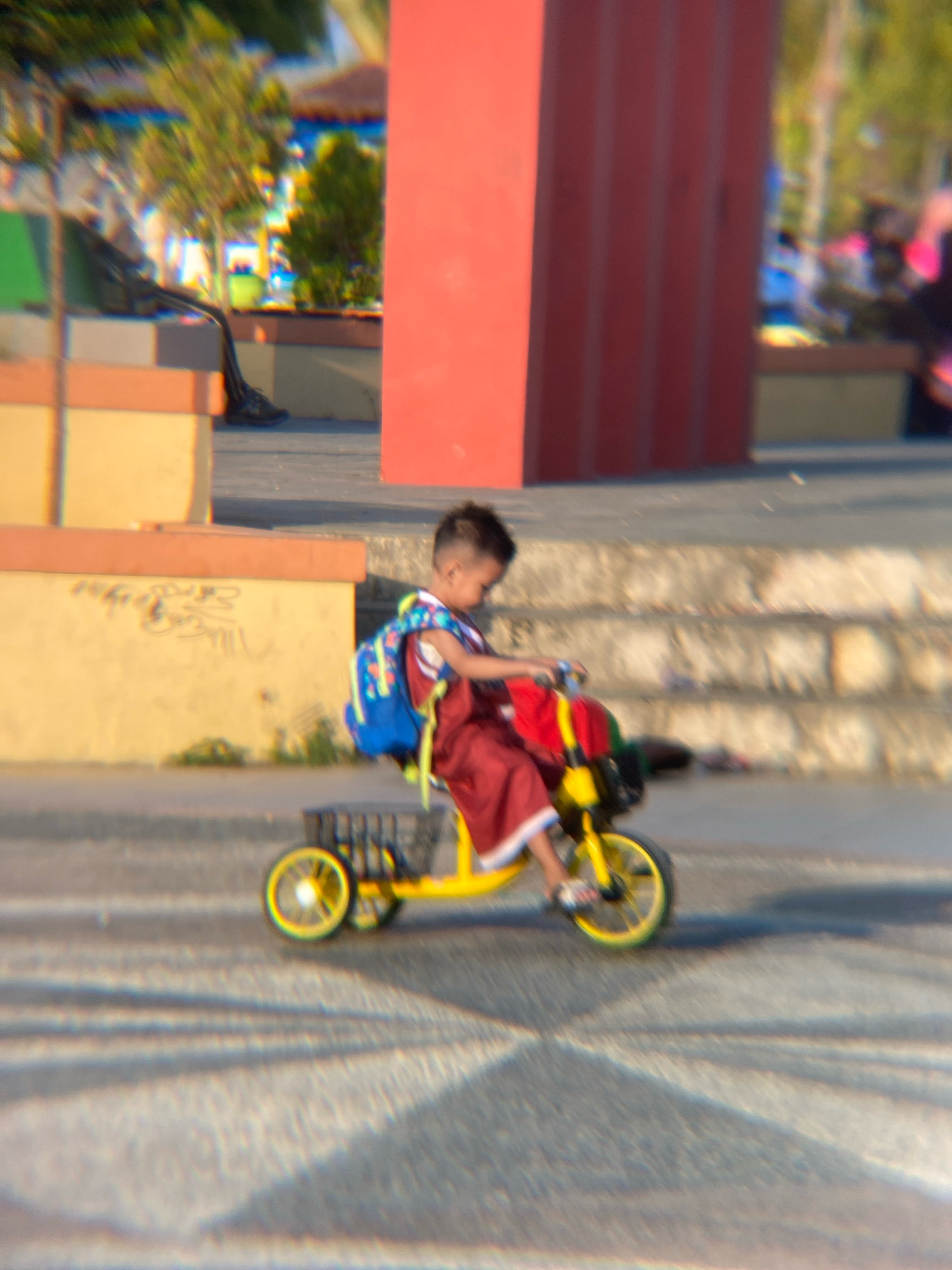 A young child rides a yellow tricycle on a paved surface with diagonal lines, possibly a road or sidewalk, in Kendari, Indonesia. The child is wearing a red garment and a blue backpack. The tricycle has a black basket on the back. In the background, a blurry red structure and stairs are visible. The overall image is out of focus, suggesting motion.