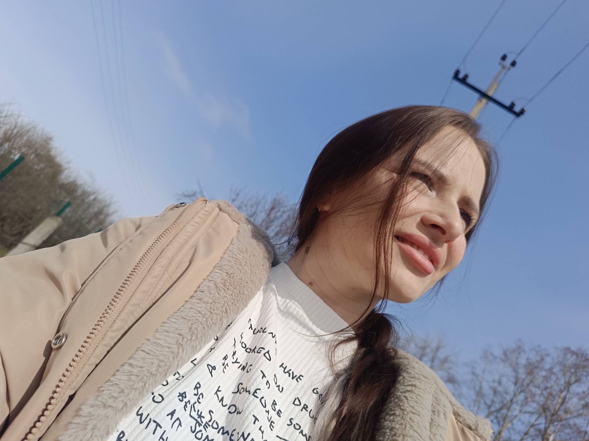 A young woman is captured in a low-angle, close-up portrait, looking upwards and to the right with a soft smile, eyes slightly narrowed as if gazing into bright sunlight. Her dark brown hair, with some strands falling across her face, is styled with a braid visible over her left shoulder. She wears a light beige or tan puffer jacket, partially open to reveal a beige faux fur lining, over a white, ribbed long-sleeved shirt. The shirt is printed with black, handwritten-style text; visible fragments include "LOOKED HAVE", "DRU", "SOMED, SE SOME", "BE", "KNOW TH", "ONE", "SON", and "WITH".
The background features a vast, clear blue sky, indicating a bright, sunny day. A utility pole with several power lines is visible in the upper right. Out-of-focus, bare tree branches are present on the left and in the lower right, suggesting a cooler season such as late winter or early spring. The strong sunlight illuminates her face and jacket. The image was taken in Фоки, Russia.