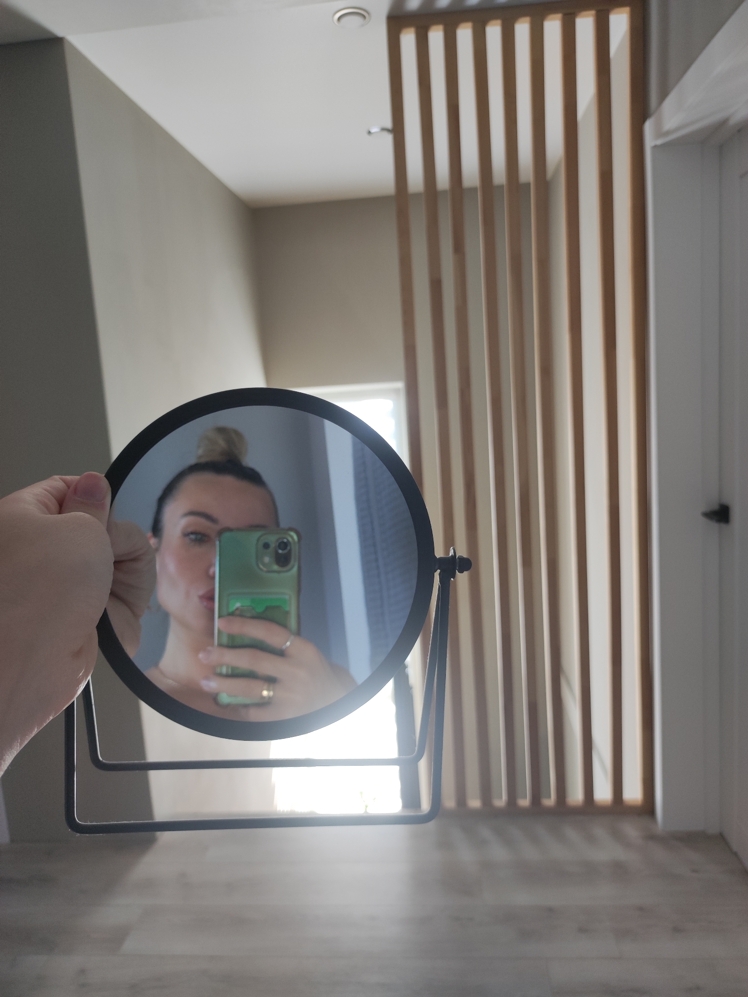 A woman is taking a selfie in a round, black-framed mirror. The woman has her hair up in a bun and is holding a green smartphone with a dual-lens camera. She is wearing a ring on her finger. The background includes a wall with vertical wooden slats, a white door, and a floor that appears to be made of light-colored wood or laminate. The location of the image in the city of Votkinsk, Russia, is unknown.