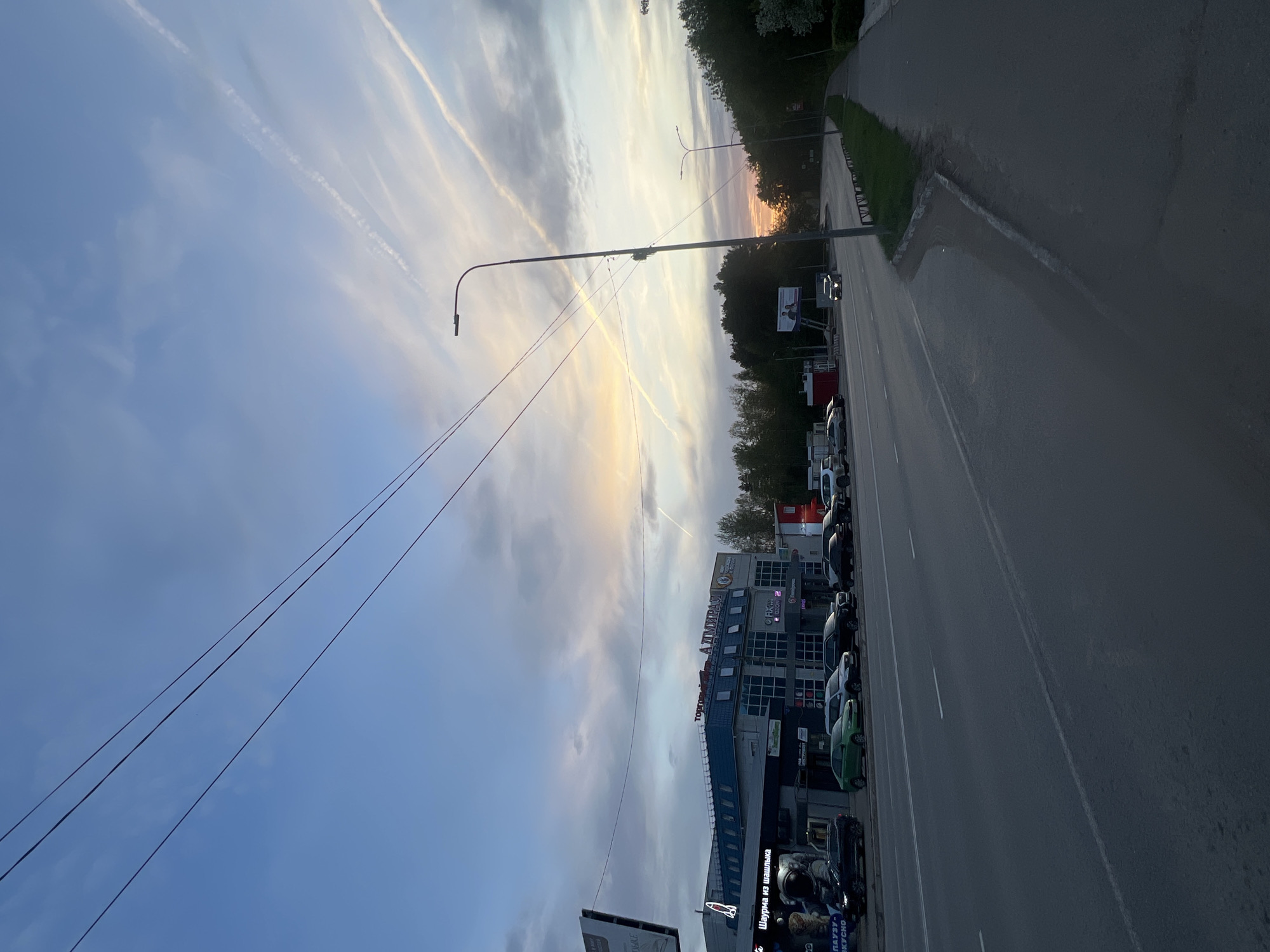 The image shows a street scene in Chaikovsky, Russia, during twilight. A multi-story building with several commercial businesses, including one that mentions "Shawarma," runs alongside the road. Cars are parked along the roadside. Streetlights illuminate the road. Contrails are visible in the sky above. A forested area is on the far side of the road. The sky transitions from blue to shades of orange and yellow near the horizon. Electrical wires stretch across the sky.