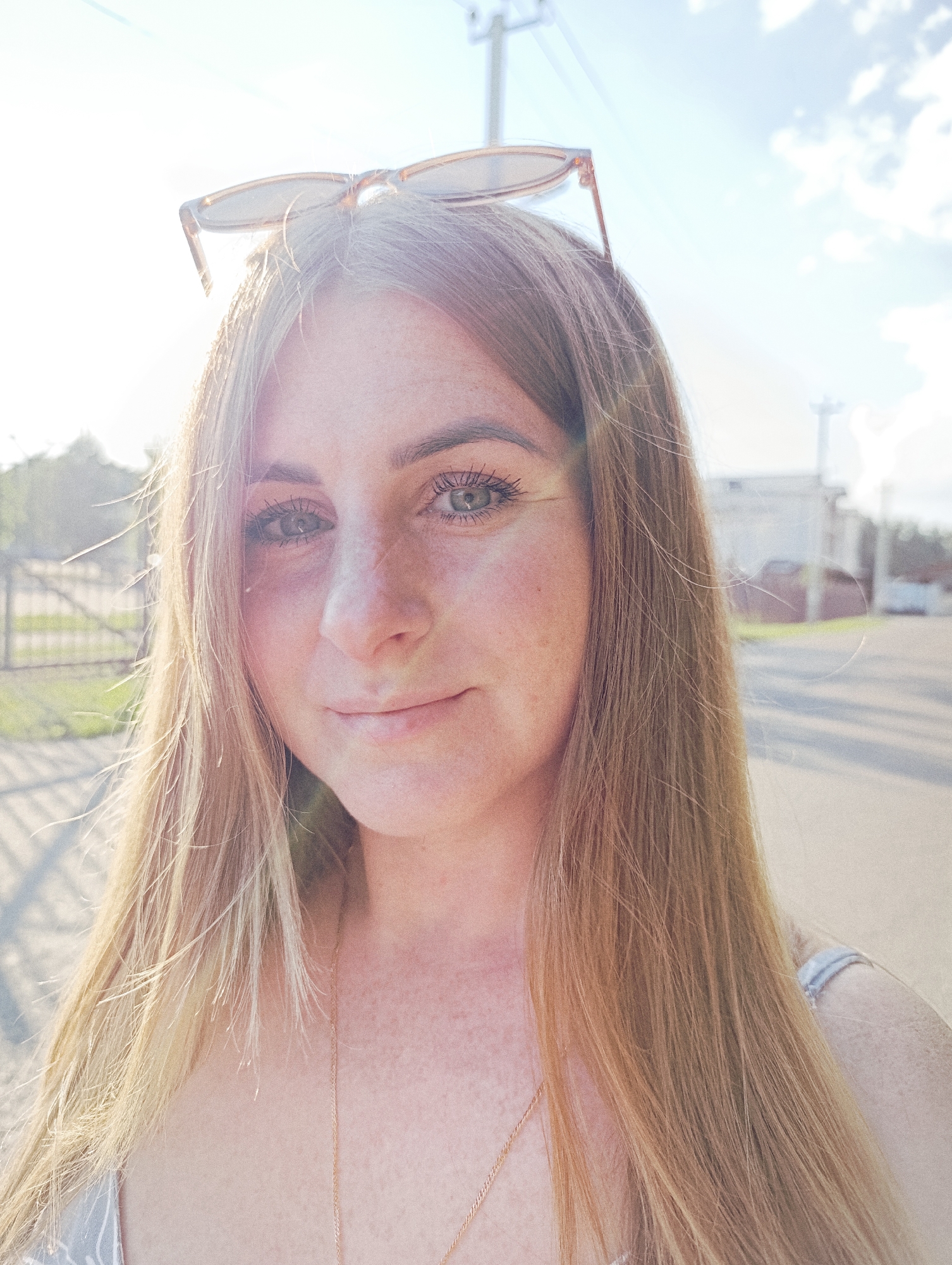 A young woman with blonde hair and freckles is shown in a close-up selfie, taken outdoors during the day, with the sun in the background creating lens flare. She is wearing sunglasses on top of her head and a necklace. The background shows a street with some buildings and a fence, suggesting an urban or suburban environment. There are clouds in the sky, indicating a partly cloudy day. The location is identified as Ivanovskoe, Russia.