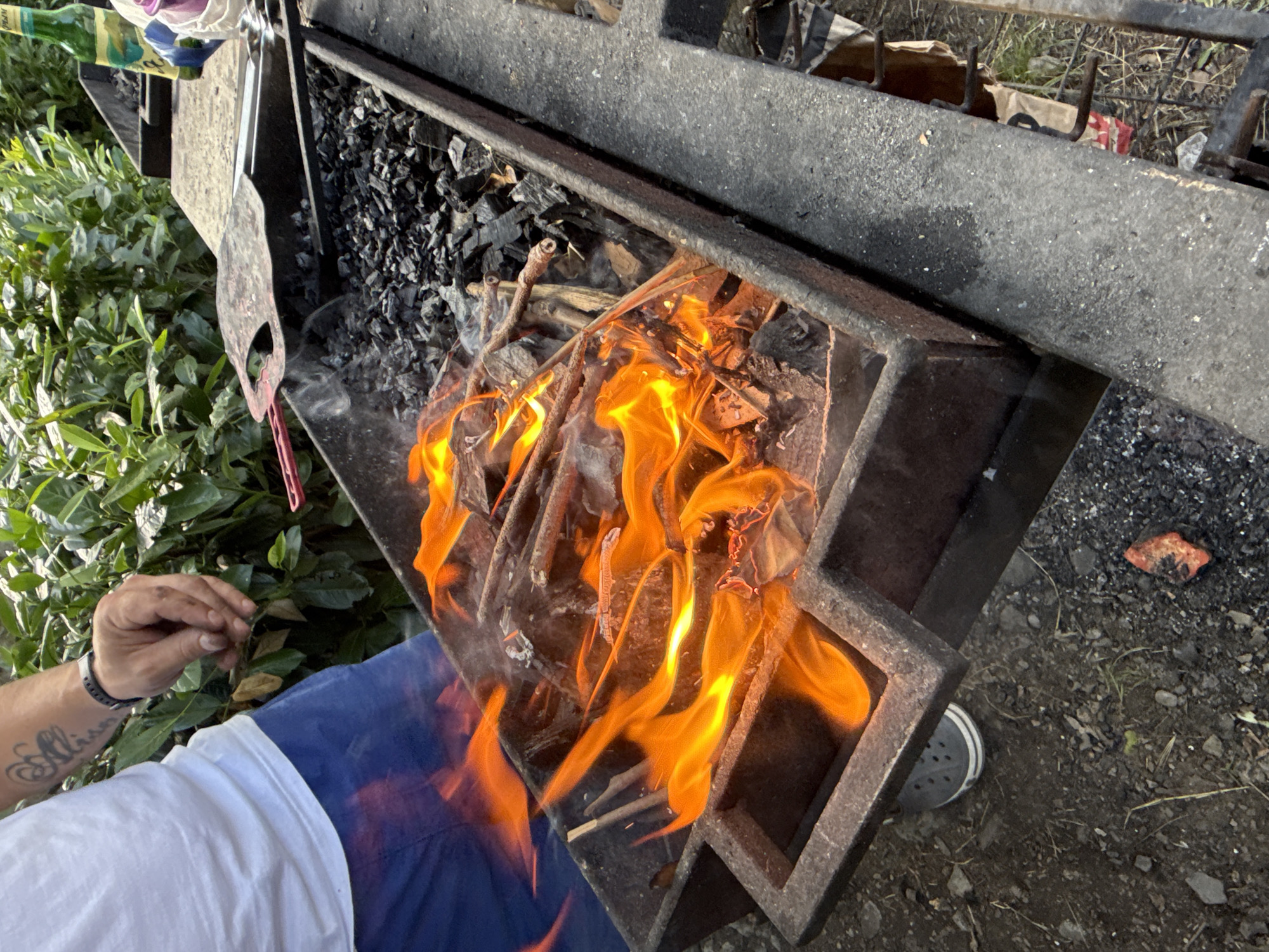 A person wearing a white t-shirt and blue shorts is standing next to a metal barbecue grill that is actively burning wood and charcoal, producing flames and smoke. The grill is rectangular with handles on the sides. A pizza peel and tongs are placed on the edge of the grill. In the background, there are green bushes and a bottle of beer. The ground around the grill is covered in dirt and pebbles. The scene appears to be outdoors. No specific location within the city of Partenit, Ukraine can be determined from the image.