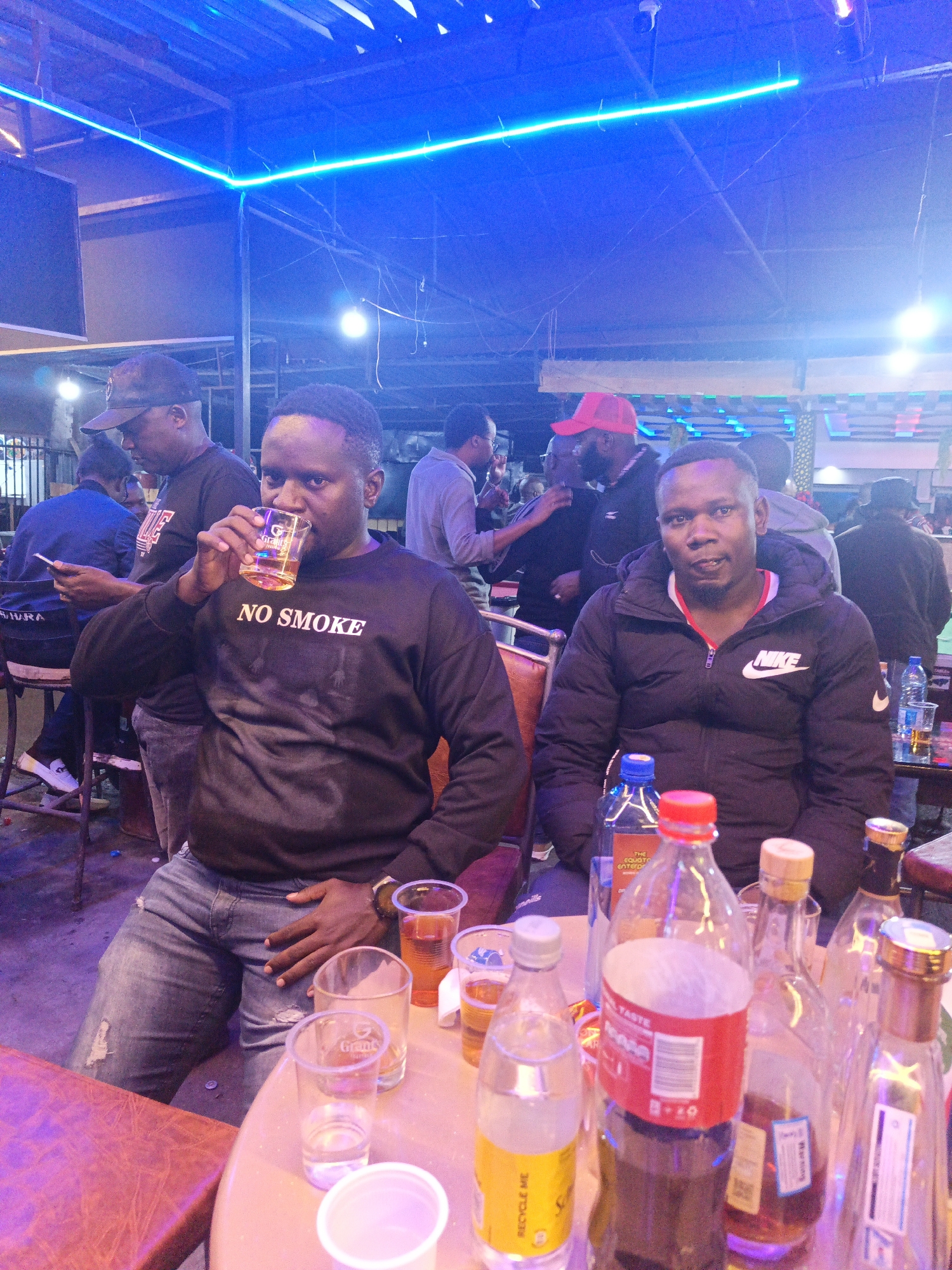 The image depicts an indoor scene, likely a bar or restaurant in Nairobi, Kenya, based on the appearance and context. Two men are prominently featured at a table filled with multiple bottles of alcohol, soft drinks, and glasses. One man wears a black sweatshirt with "NO SMOKE" printed on it and is in the process of drinking from a glass. The other man wears a black Nike jacket. Other individuals are present in the background, some seated at a bar. The lighting is dominated by a bright blue neon light fixture above the table, giving the scene a slightly artificial, stylized appearance. The general atmosphere suggests a social gathering.