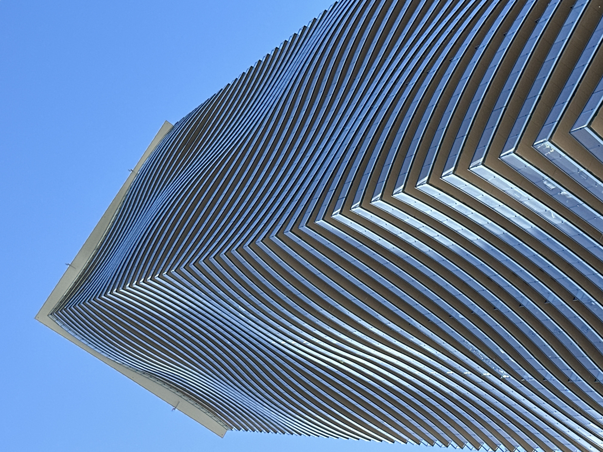 This is a low-angle, full shot of the curved facade of a modern skyscraper in Burnaby, Canada. The building has a distinctive wavy design, with floors appearing to undulate as they ascend. The exterior is primarily composed of glass balconies and horizontal metallic fins. The sky is a clear, bright blue, indicating a sunny day. There are no people or other visible objects in the frame.