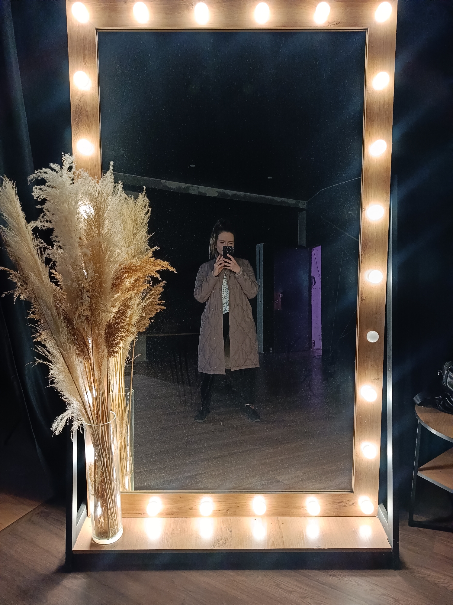 A full-length photograph depicts a person taking a mirror selfie. The individual is centrally positioned in the mirror's reflection, wearing a light brown quilted coat over patterned clothing and dark pants. They are holding a smartphone up to take the picture.
The mirror itself is a large, rectangular vanity mirror with an illuminated wooden frame. A series of light bulbs line the periphery of the frame. To the left of the mirror is a large glass vase containing decorative dried plants or reeds.
The setting appears to be indoors, possibly a studio or dressing room. Behind the person in the reflection, a dark door is visible, along with blurred interior details. The background immediately around the mirror is dark, suggesting a studio environment or a minimally lit space. To the right of the mirror, a small round table with a tiered shelf can be seen, upon which an item resembling a bag or accessory is placed. The floor is wooden.
Based on the metadata and user prompts, the location is Chaikovsky, Russia.
