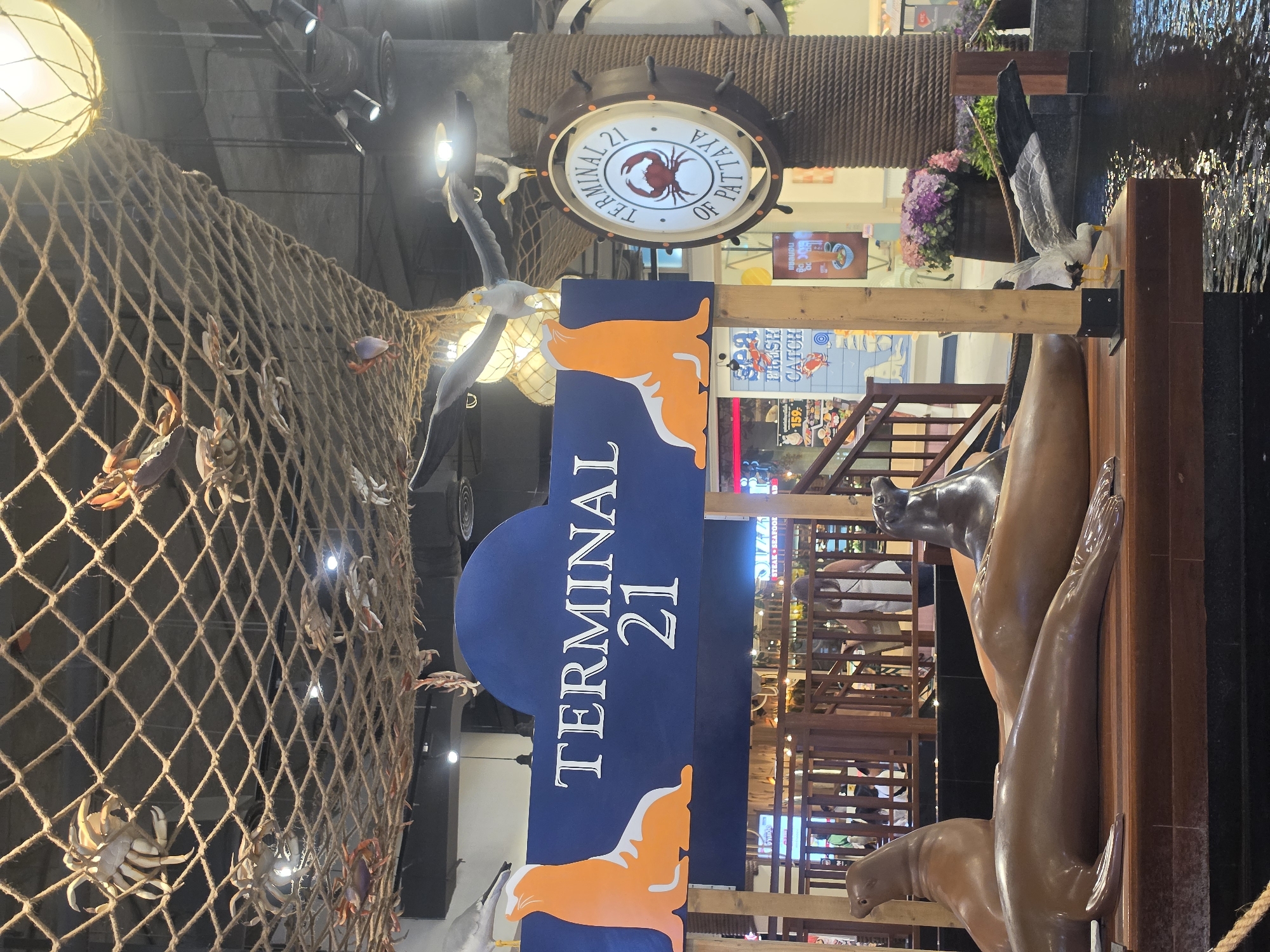 The image shows an interior scene at Terminal 21 Pattaya, a shopping mall in Pattaya City, Thailand. A prominent feature is a sign that reads "TERMINAL 21," with orange wave-like graphics at the top and bottom. Beside the sign are sculptures of seals. To the left of the sign is a large fishing net, adorned with plastic crabs and lobsters. Above the sign, a sign shaped like a ship's wheel indicates "TERMINAL 21 OF PATTAYA" with a crab emblem in the center. Faux seagulls are scattered throughout the display. Shoppers are visible in the background.