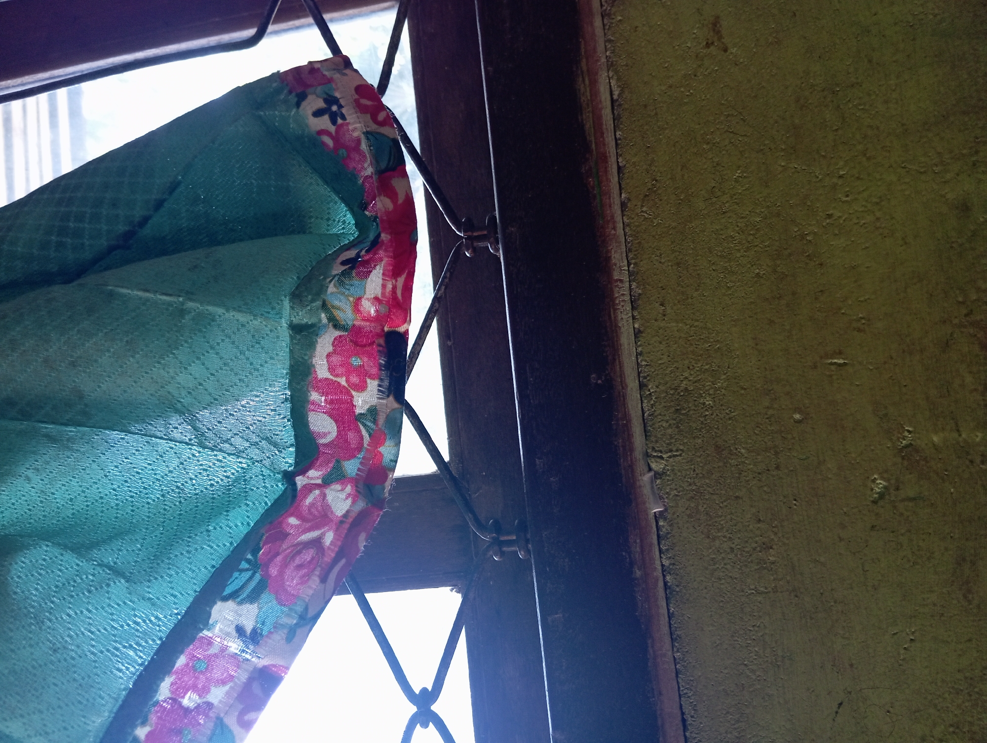 A close-up, vertical shot captures a section of a window in Rejosari, Indonesia. A teal-colored fabric, possibly a curtain or part of a garment, is draped across the top, featuring a floral trim with pink and white blossoms. The fabric is textured and appears somewhat damp. The window is fitted with a dark metal grate, its bars forming a diamond pattern. Below the window, a dark wooden frame is visible, and beneath that, a roughly textured, olive-green wall. The overall lighting suggests it is daytime, with light filtering through the window. No people or specific activities are depicted.