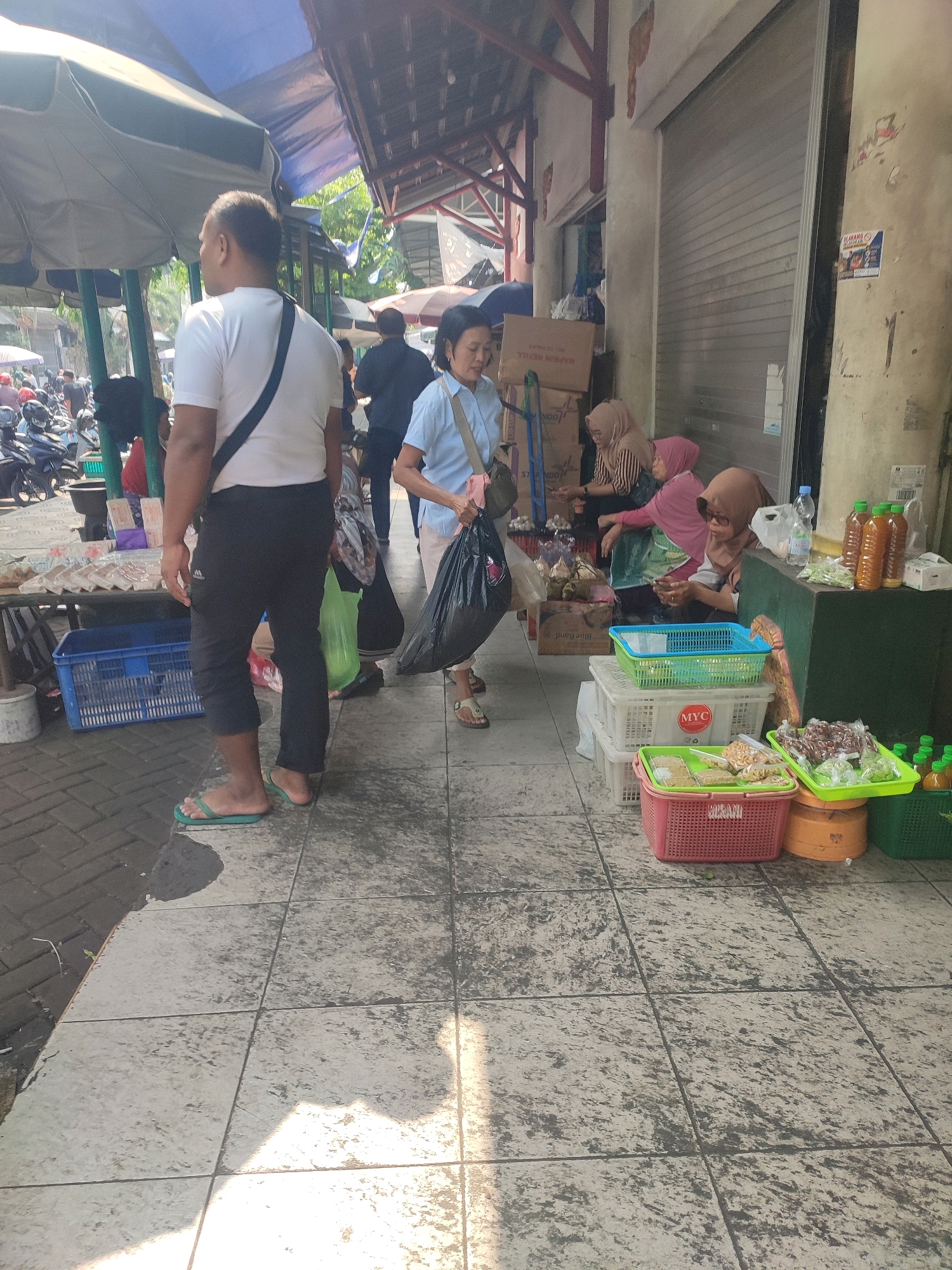 The image captures a scene in a market in Yogyakarta, Indonesia, likely on a sunny day, with strong shadows indicating mid-day. In the foreground, a man wearing a white t-shirt, black trousers, and green flip-flops walks away from the camera. He carries a black bag over his right shoulder. To his right, a woman in a light blue shirt and light-colored pants, also carrying a black bag, is walking away from the camera. She is in the middle ground, slightly further from the viewer than the man.
Behind them, on the right side of the image, a stall is set up with various items for sale. Three women are visible at this stall. One, wearing a pink hijab and a pink garment, is seated and looking at her phone. Another woman, wearing a striped top and a light brown hijab, is also seated. A third woman, wearing a brown hijab and a gray patterned top, is partially visible, also seated. The stall displays several plastic baskets filled with goods. One red basket contains packaged snacks, and a green basket holds more snacks or food items. There are also bottles of liquid, possibly beverages, and some green vegetables or fruits.
The environment appears to be an open-air market or a street with stalls. A corrugated metal shutter door is visible behind the stall. The ground is paved with square tiles, and there's a slight wetness on the pavement in some areas. Various motorcycles are parked in the background on the left, suggesting a busy area. Overhanging tarps and awnings provide shade. The overall impression is of a bustling, everyday market scene in an Indonesian city.