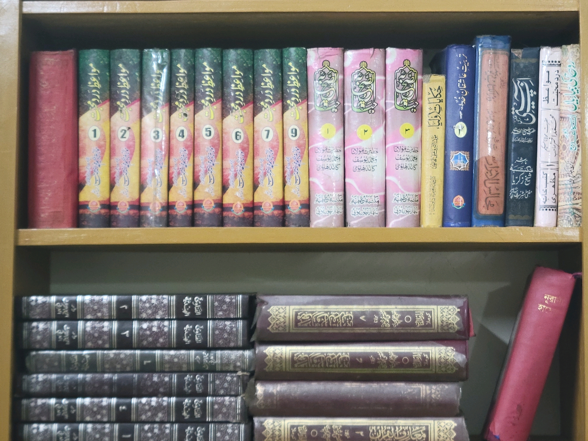 The image shows a wooden bookshelf filled with books. The top shelf has several books with colorful spines, some featuring Arabic calligraphy and numbering. The spines are predominantly green, pink, yellow, and blue. A red book is positioned on the far left of this shelf. The bottom shelf displays a stack of books with dark covers and gold lettering. On the far right of the bottom shelf, a single red book is standing upright. The environment appears to be an indoor setting, likely a home or library, with the bookshelf as the primary focus. There are no people or animals visible in the image. The city of Sindurpur, Bangladesh, is not discernible from the visual information provided in the image.