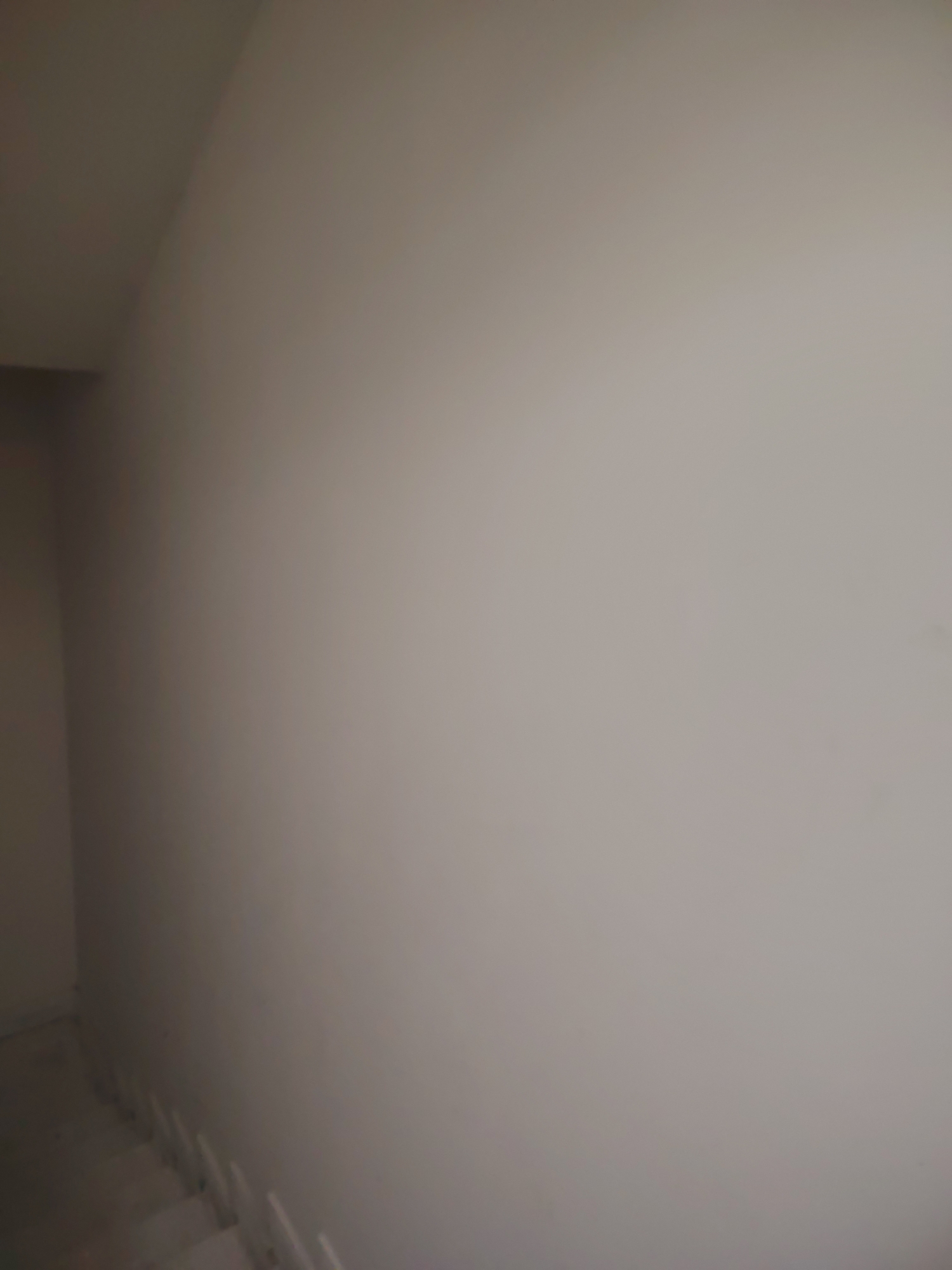 The image displays an interior architectural scene, primarily focusing on a plain wall and a section of a staircase. The main subject is a large, unadorned wall, which occupies the majority of the frame. This wall appears to be painted in a neutral, light gray or beige color, with subtle variations in shading that suggest gentle, diffuse lighting.
In the upper left corner, an angled boundary indicates a ceiling or an adjacent wall, creating a corner within the room. Along the bottom and extending towards the lower left, several steps of a staircase are visible, ascending or descending. These steps are also light-colored, matching the overall subdued palette of the space, and appear to be simple in design, possibly unfinished or part of a minimalist aesthetic.
No people are present in the image, and no specific activity or event is taking place, giving the scene a static and quiet quality. The lighting is soft and even, making it difficult to determine the time of day, though it appears to be an interior setting, possibly with indirect natural light or artificial illumination. There are no identifiable location cues beyond a generic indoor stairwell or hallway, and no visible text or explicit emotions conveyed. The overall impression is one of starkness and simplicity.