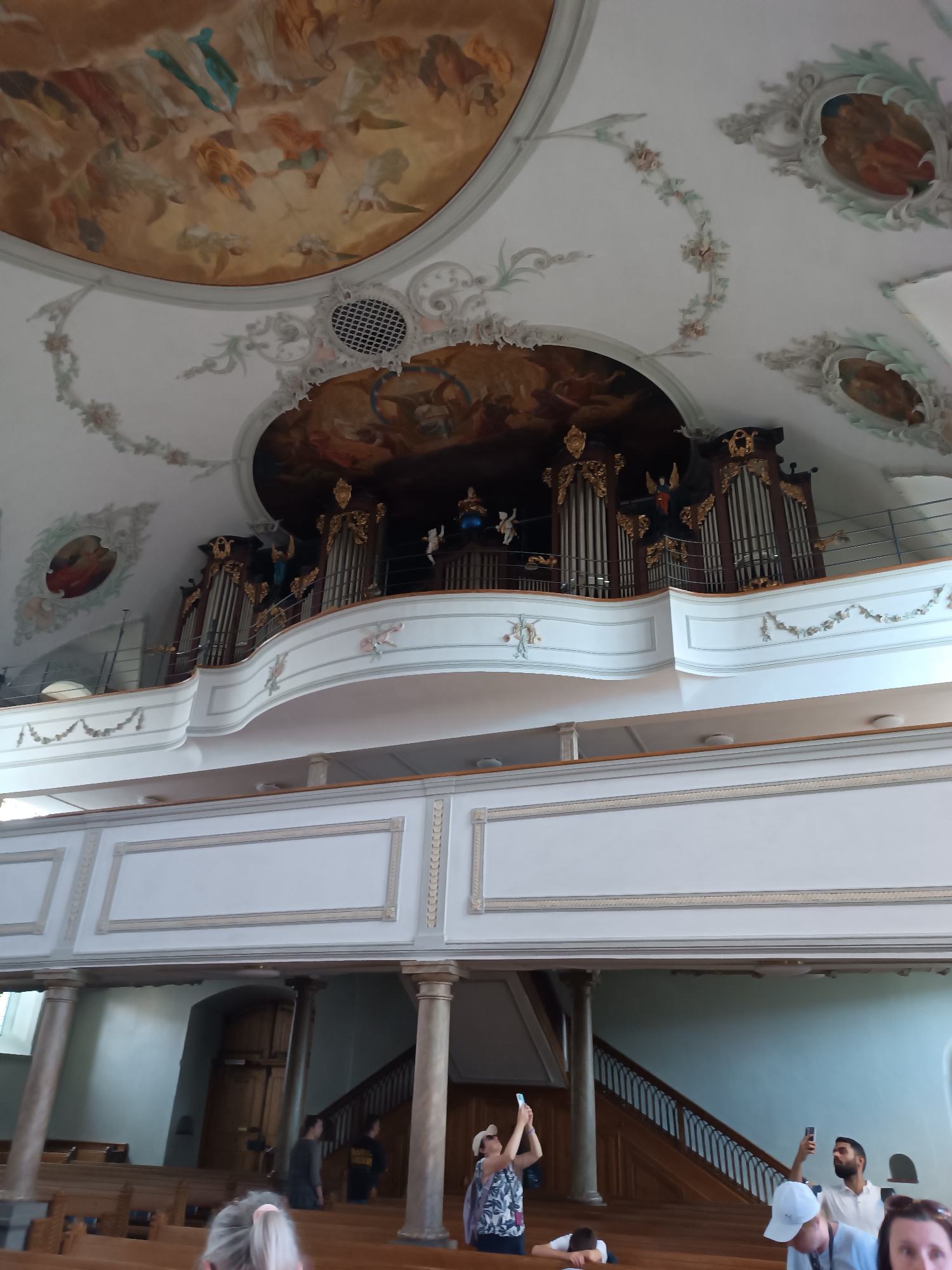 The image depicts the interior of a church, likely the Appenzell Catholic parish church, St. Mauritius, in Appenzell, Switzerland. The ornate Baroque ceiling is adorned with frescoes of religious scenes, featuring cherubs and figures. Below the ceiling, a grand organ with intricate wooden carvings and golden accents is situated on a balcony. The balcony itself is white with decorative trim and is supported by pillars. In the foreground, rows of wooden pews are visible. Several individuals are present, some standing and observing, while others are seated. One person in the mid-ground is taking a photograph with their phone. Another person in the foreground, partially visible, also appears to be holding a phone. The overall ambiance suggests a place of worship and historical significance.