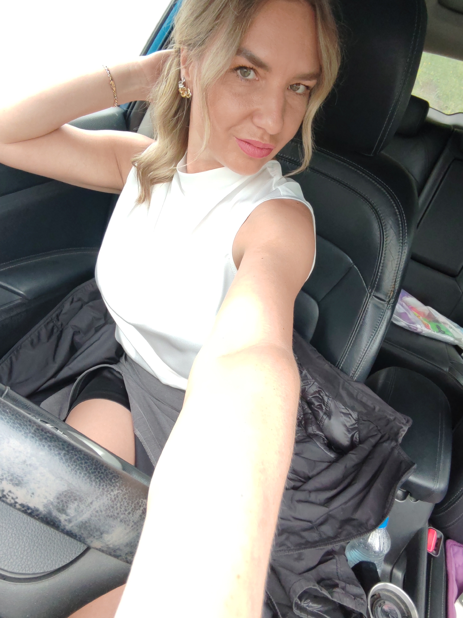 A woman with blonde hair and light skin is in the driver's seat of a car. She is wearing a white sleeveless top, a gray skirt, and black shorts. Her right arm is raised, and she is touching her hair with her hand. She is wearing gold earrings and a gold bracelet. Her makeup includes pink lipstick and eyeliner. The car has black leather seats. A black jacket is draped over the passenger seat, and a bottle of water is in the center console. The background is blurred, but appears to be an outdoor scene with greenery.