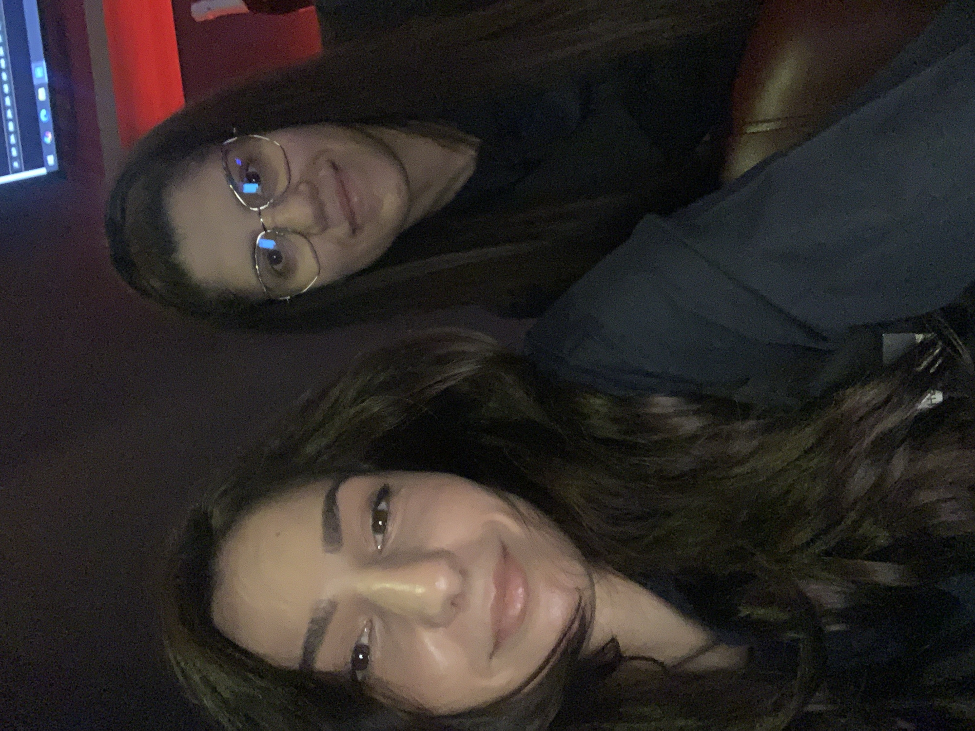 The image shows a selfie taken by two young women indoors. The woman in the foreground has long, dark, wavy hair and is looking directly at the camera with a slight smile. The woman in the background, wearing glasses, is also looking at the camera and smiling. They appear to be in a dimly lit room, possibly a studio or entertainment venue, with a computer monitor visible in the upper right corner and red lighting in the background. There are no visible signs of the city of Burgas, Bulgaria, in the image.