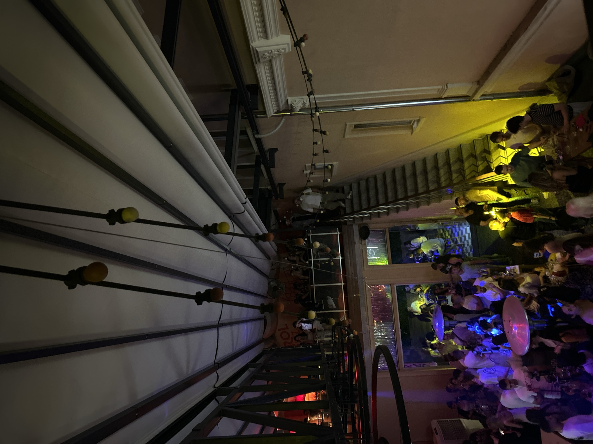 This image captures a lively scene at a bar in Plovdiv, Bulgaria. The setting is a partially covered outdoor area with many people. Tables are filled with customers who appear to be eating, drinking, and socializing. String lights and architectural details adorn the ceiling. The crowd is diverse, with groups of people engaged in conversation and enjoying the atmosphere. The bar has a visible window and an open entryway. A staircase is present, leading upwards. The scene is illuminated by a combination of natural and artificial light, creating a warm and inviting ambiance.