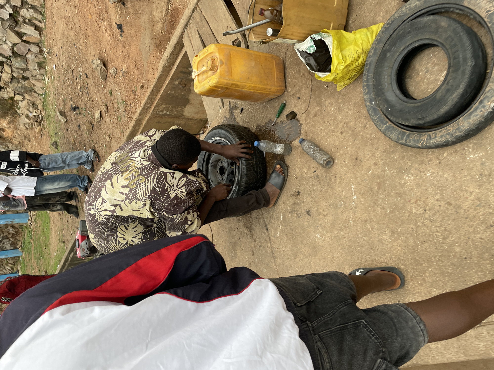 The image depicts an outdoor scene, possibly in the city of Jos, Nigeria, with individuals engaged in what appears to be tire repair work. The foreground is dominated by the lower half of a person wearing dark denim shorts and gray sandals, partially obscuring the ground. To their right, a man in a patterned shirt is bent over, working on a tire. The ground is a mix of concrete and packed earth, with scattered debris.
Key objects in the scene include:
* **Tires:** At least two tires are visible, one being worked on and another lying flat on the ground.
* **Containers:** A large yellow plastic jerrycan and a yellow bucket are present, containing various items, including a bottle and what looks like a tool.
* **Tools:** A green-handled tool (likely a screwdriver or similar implement) and a metal rod are visible.
* **Water bottles:** Two plastic water bottles are on the ground.
* **Debris:** Scattered rocks, dirt, and some black plastic bags are present.
* **Clothing:** The individuals are wearing casual clothing, including jeans, a patterned shirt, and a jacket.
The overall impression is one of a practical, possibly roadside, repair activity in an urban or semi-urban environment. The background shows a raised concrete structure and a rocky embankment, suggesting a somewhat uneven terrain.
