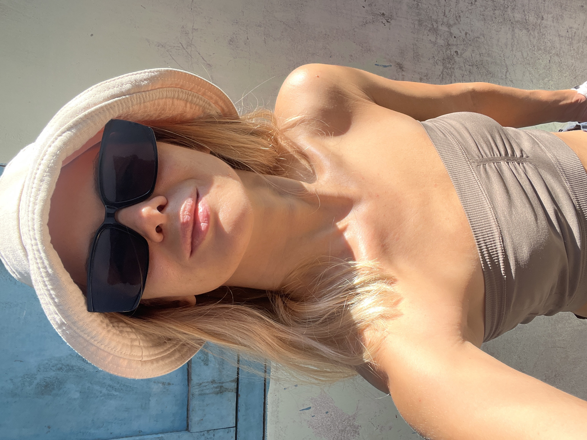 A close-up selfie shows a young woman wearing a light-colored bucket hat and dark sunglasses, with her head tilted slightly. She is also wearing a brown strapless top. Her blonde hair is visible around her face. The background consists of a textured wall in a neutral color and a section of a blue surface, possibly a door or window shutter, on the left. Sunlight casts shadows on the wall and illuminates the woman's skin, highlighting her shoulders and neck. The scene appears to be outdoors, with the bright sunlight suggesting a warm climate. It is impossible to determine the specific location in Nha Trang Ward, Vietnam, from the visual information provided.
