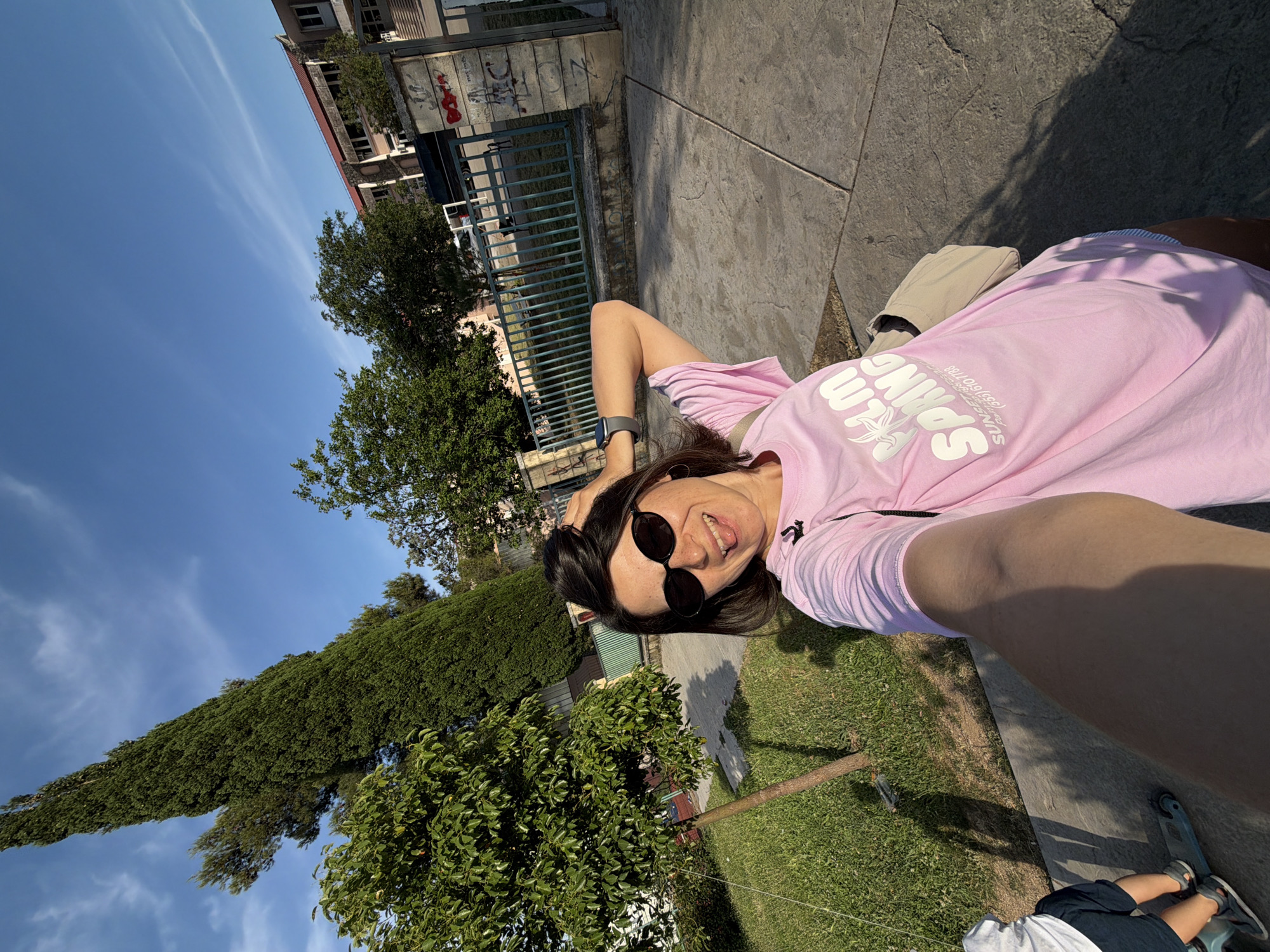 A woman in sunglasses and a pink t-shirt lies on a concrete surface. Behind her is a grassy area with trees. In the background are buildings with graffiti on the wall and a blue fence. The sky is blue with some clouds. The photo appears to be taken in Budva, Montenegro. A child sits at the bottom of the frame.