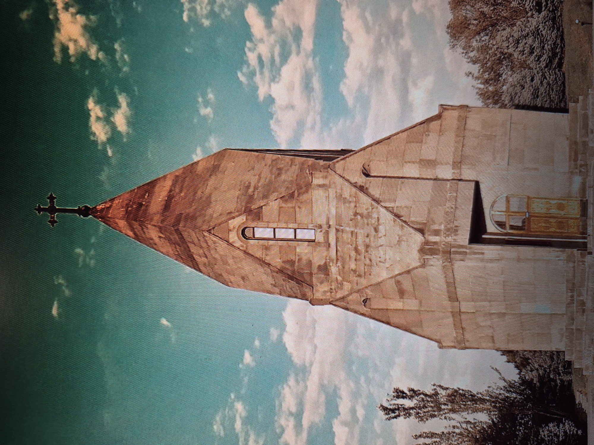 The image depicts the Holy Mother of God Church (Katoghike Church), located in Yerevan, Armenia. The church is captured from a low-angle perspective, showcasing its unique architectural style with a tall, cone-shaped roof topped with a cross. The church's exterior appears to be made of light-colored stone. A horizontal window is visible on the roof's front facade. The sky forms the backdrop, displaying a mix of turquoise and white clouds. A tree is visible on the right, with branches extending towards the church. The image has a slightly rotated orientation.