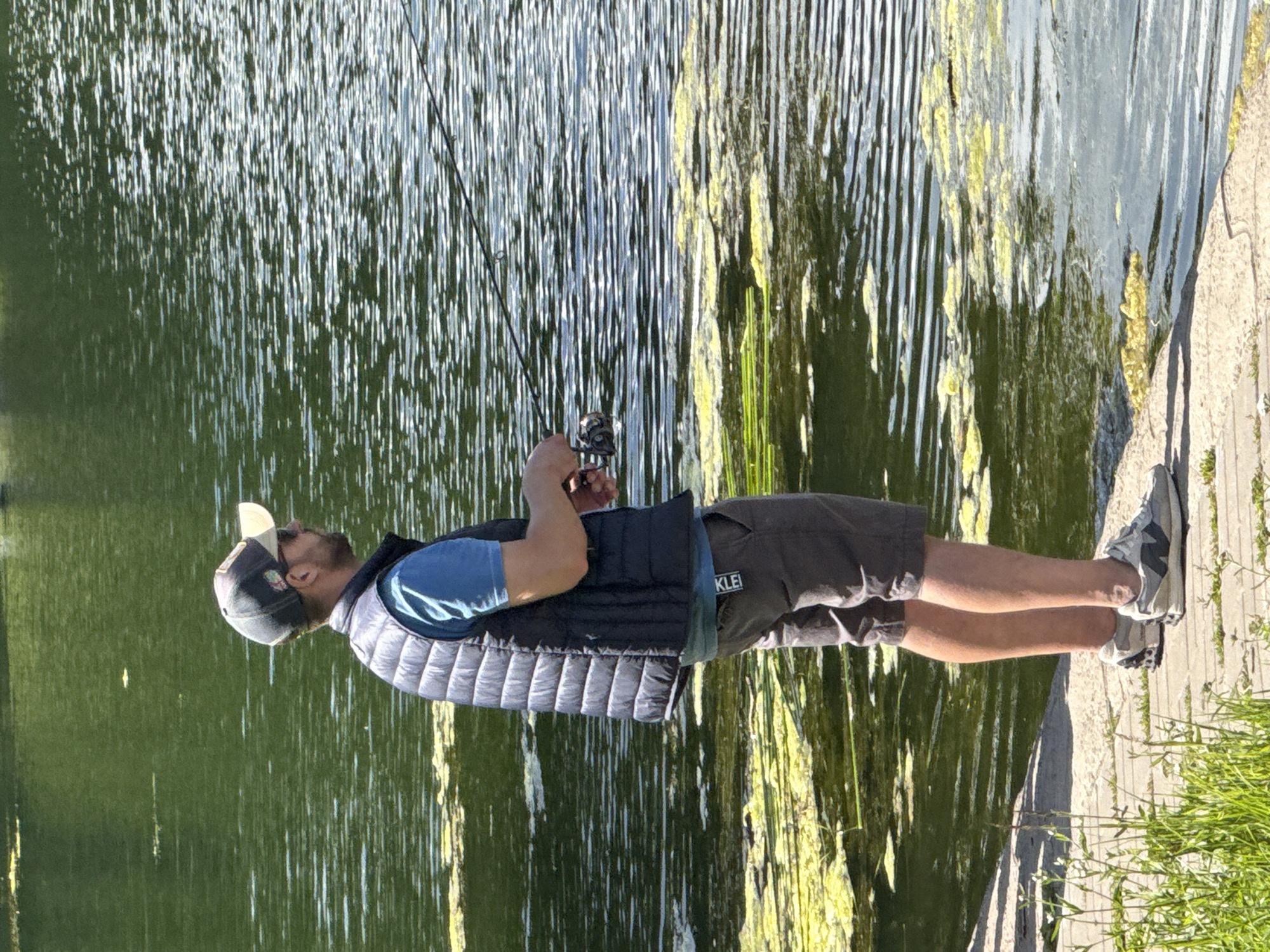An adult male is fishing on a wooden pier by a lake in a park. He is wearing a blue t-shirt, a dark puffer vest, grey shorts, a baseball cap, sunglasses, and athletic shoes. He is holding two fishing rods. The lake water is dark green with patches of yellow algae on the surface. The pier is made of concrete slabs and there is grass and small white flowers growing around it. The background is blurred with green foliage. The specific location within Moscow, Russia, cannot be determined from the image alone.